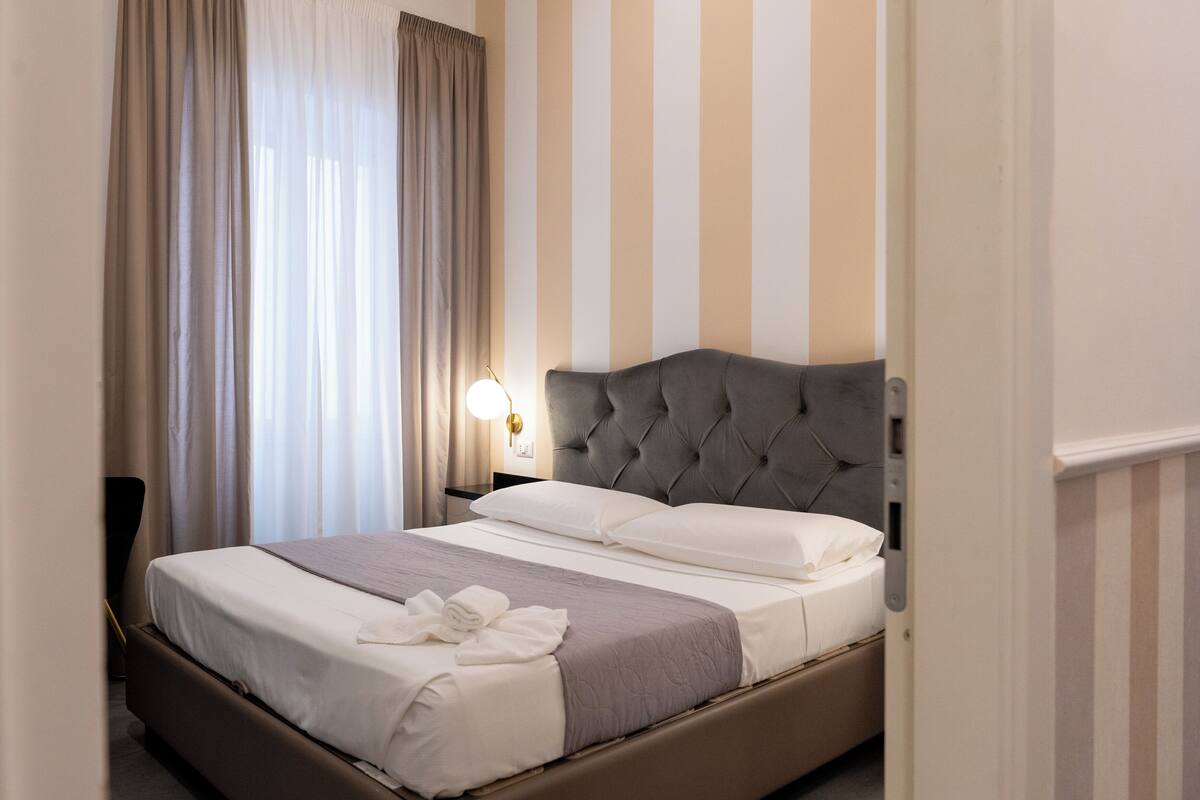 A neatly made bed with a grey headboard, white linens, and a grey blanket. There is a bedside lamp on a nightstand.