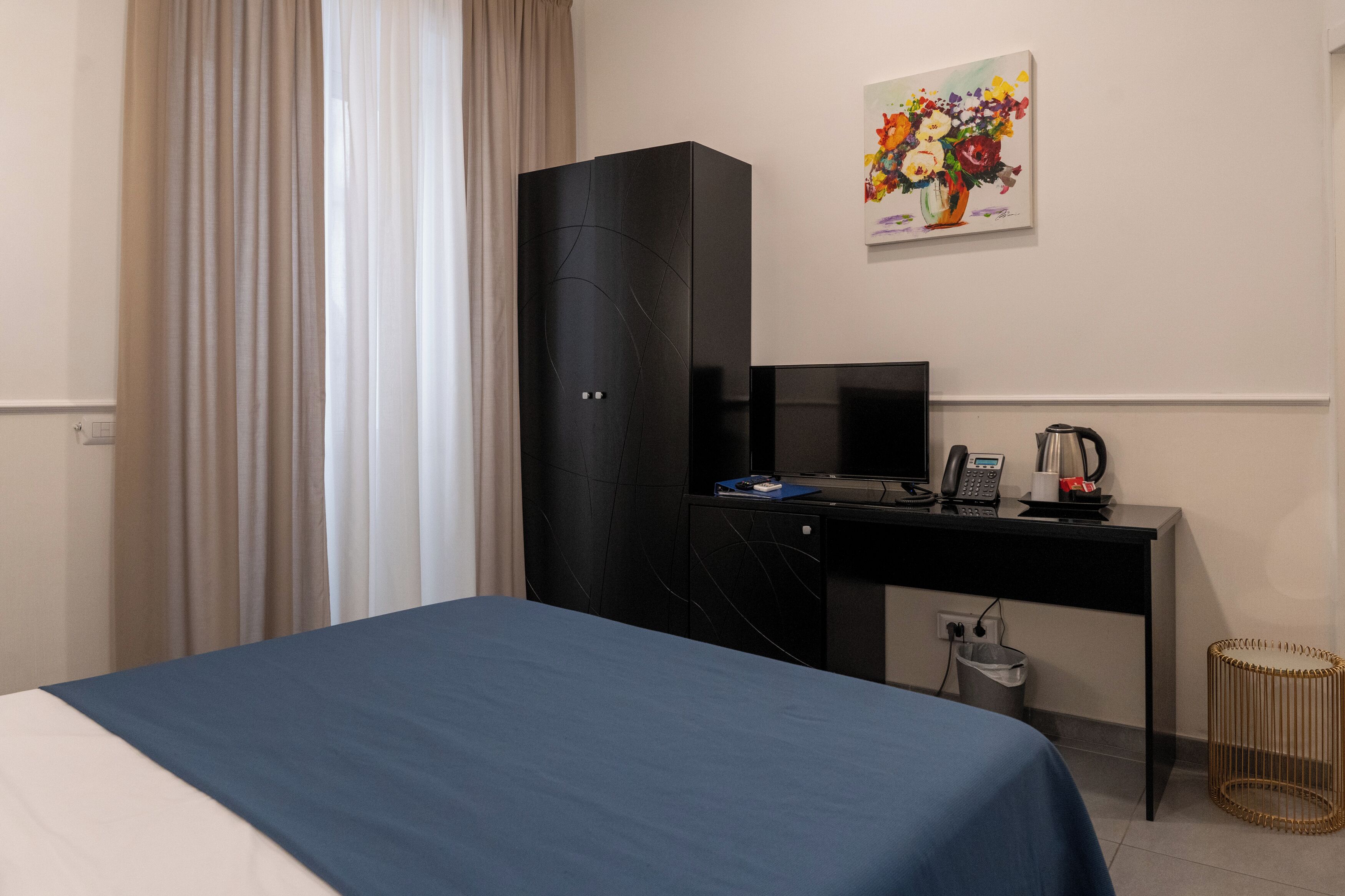 Een hotelkamer met een bed, een bureau met computer en telefoon, een prullenbak en een schilderij aan de muur.
