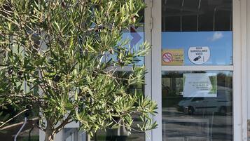 Entrada de un edificio con un árbol en maceta, una cámara de seguridad y una puerta con un cartel que indica vigilancia y un letrero de estacionamiento.