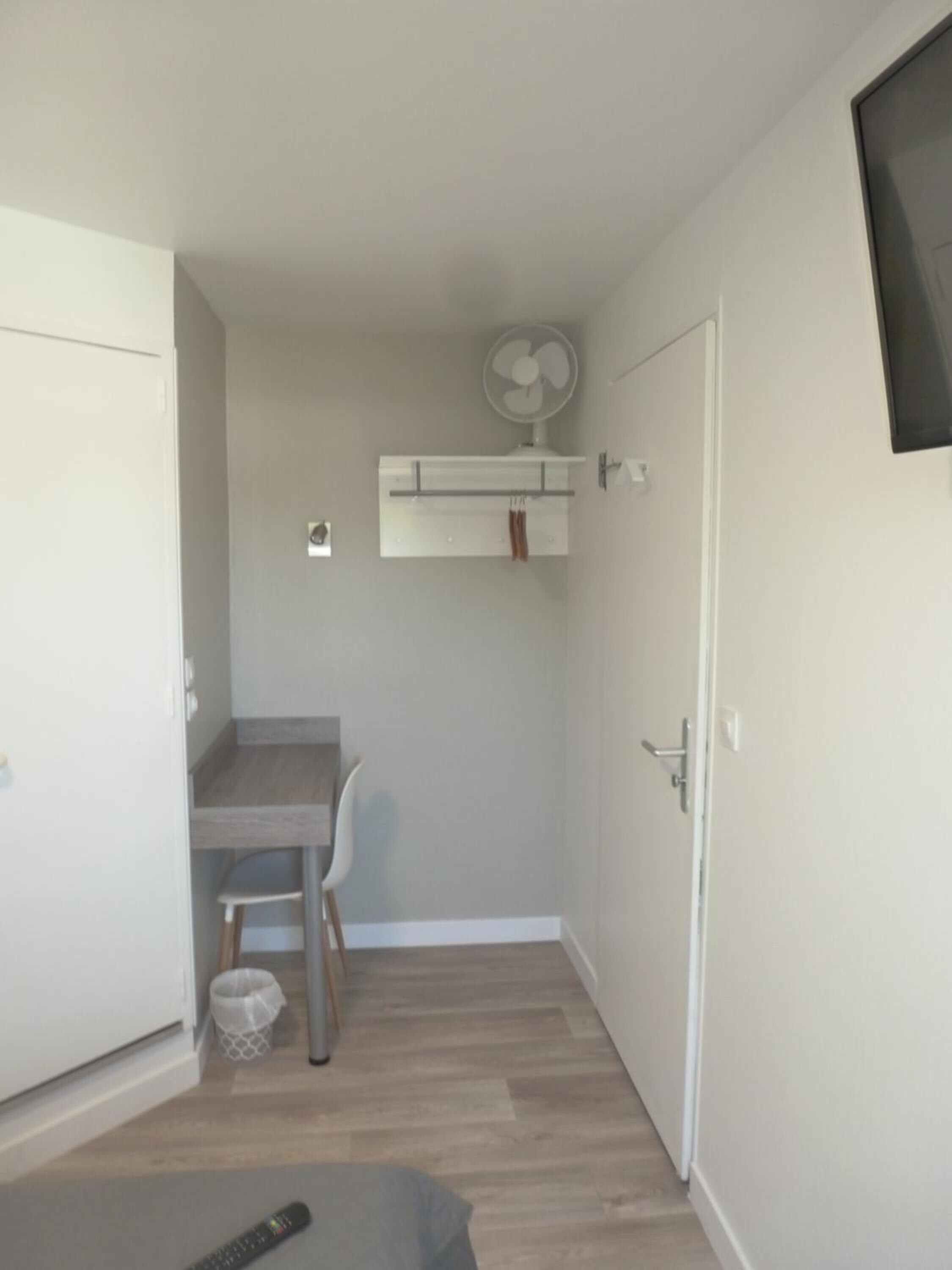 Een kleine, praktische kamer met een bureau, stoel en een ventilator aan de muur.