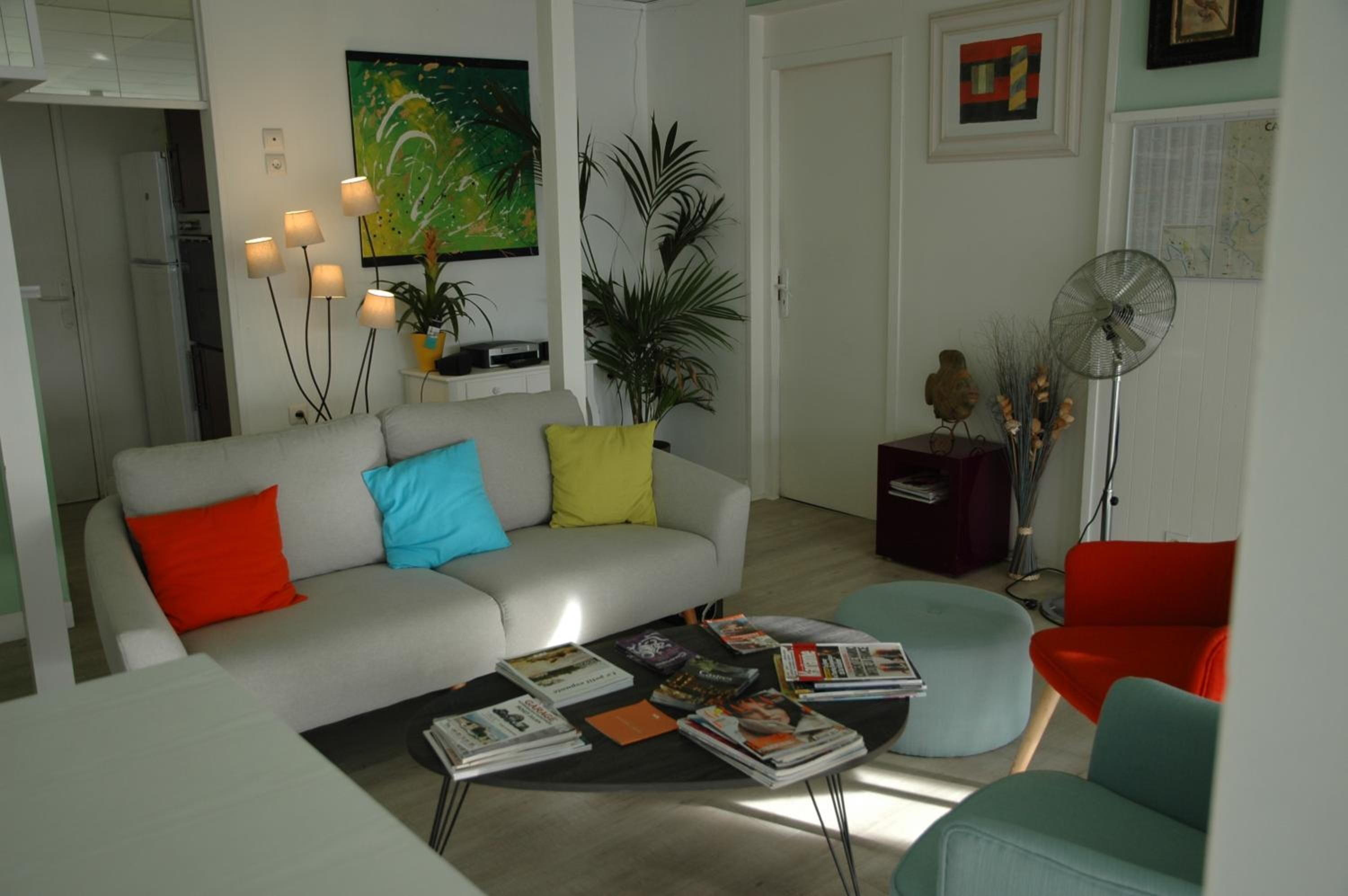 A living room with a sofa, ottoman, and a coffee table with magazines. There is a fan and a plant in the room.