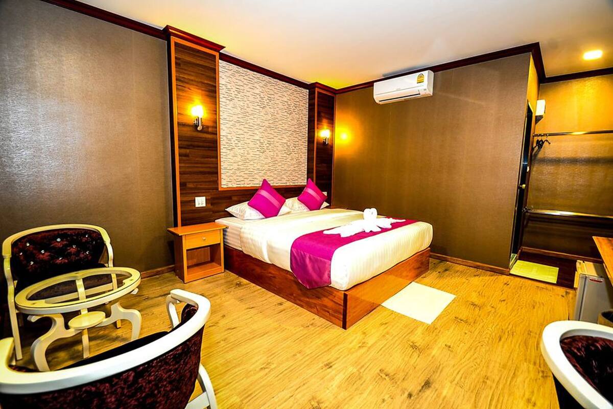 Habitación de hotel con cama, una silla, una mesita, una mesita de noche, un equipo de aire acondicionado y suelo de madera.