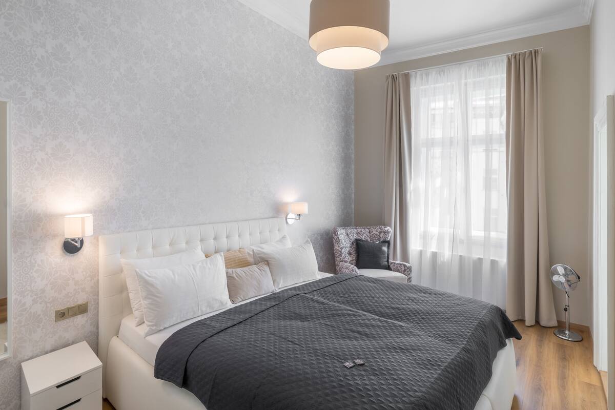 A modern bedroom with a tufted headboard, a large bed, bedside lamps, a chair, a standing fan, and sheer curtains.