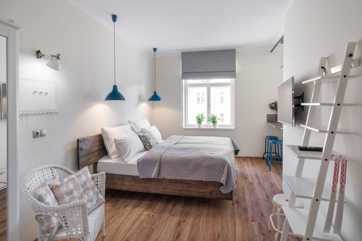 A bedroom with a wooden bed, white walls, a window with blinds, a blue pendant light, a white chair, and a white ladder.