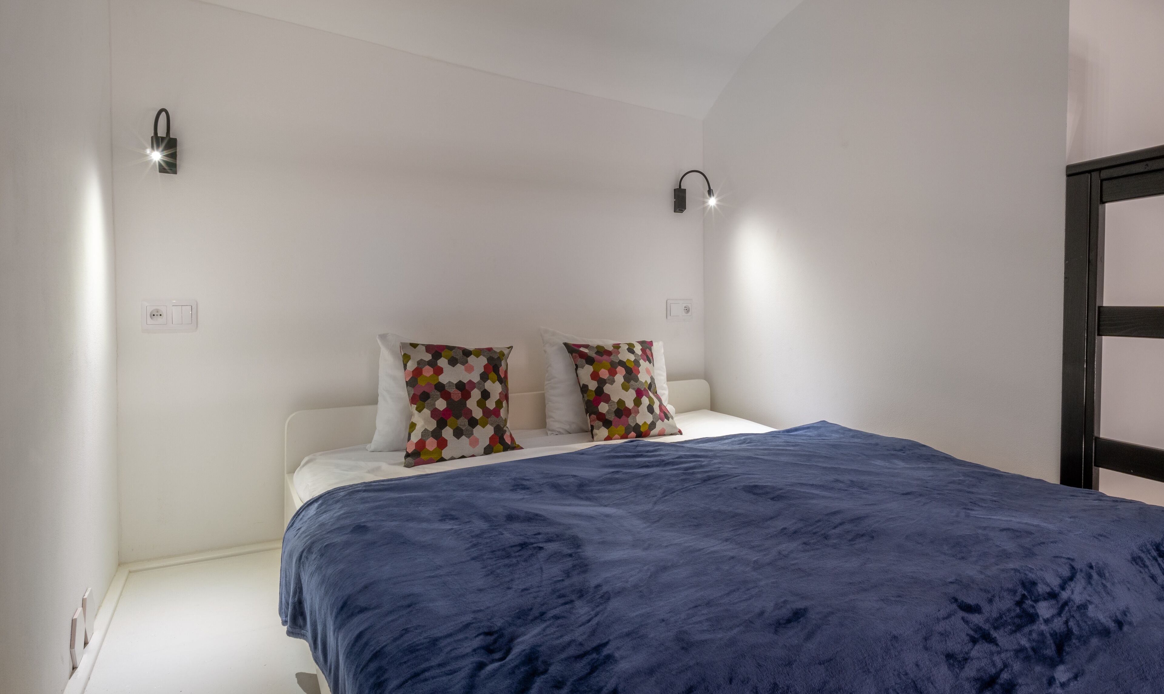 A bedroom with a bed, two patterned pillows, a blue blanket, a wall-mounted light, and a power outlet.