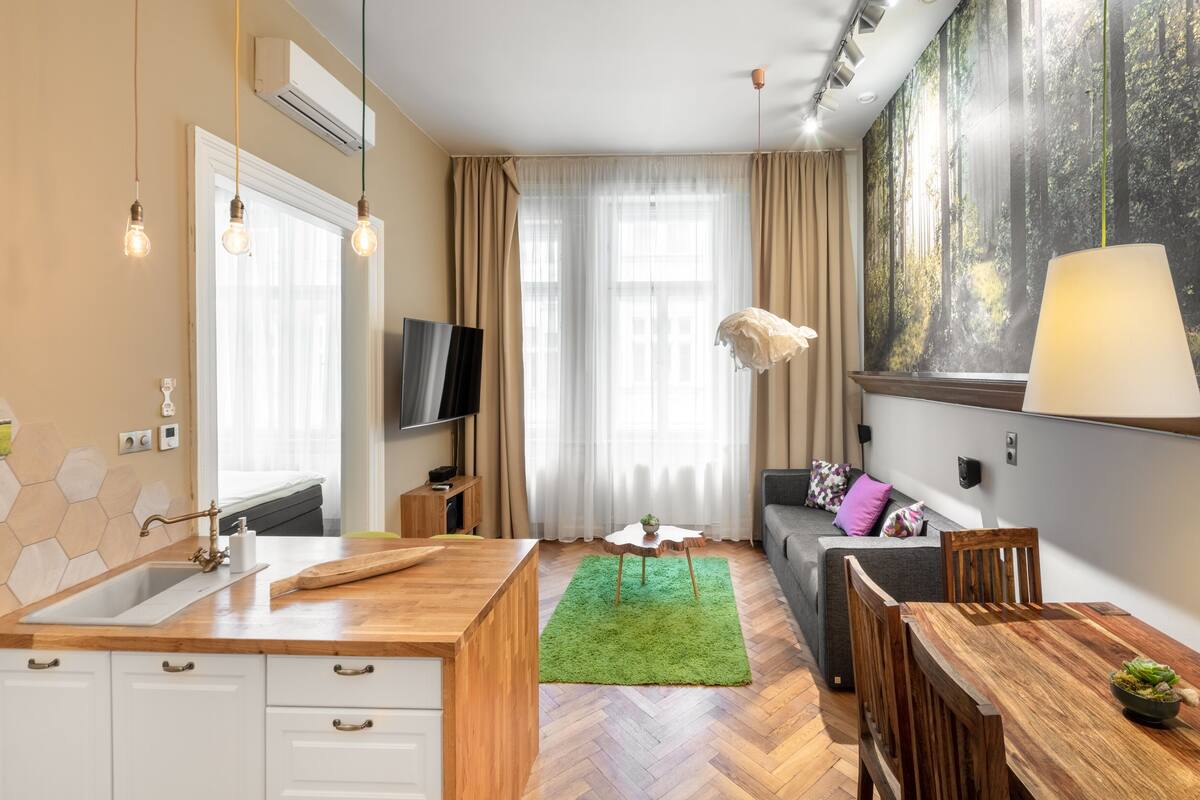 A modern kitchen with a wooden countertop, a sink, and a dining area with a table and chairs. There is a TV mounted on the wall and a large window with curtains.