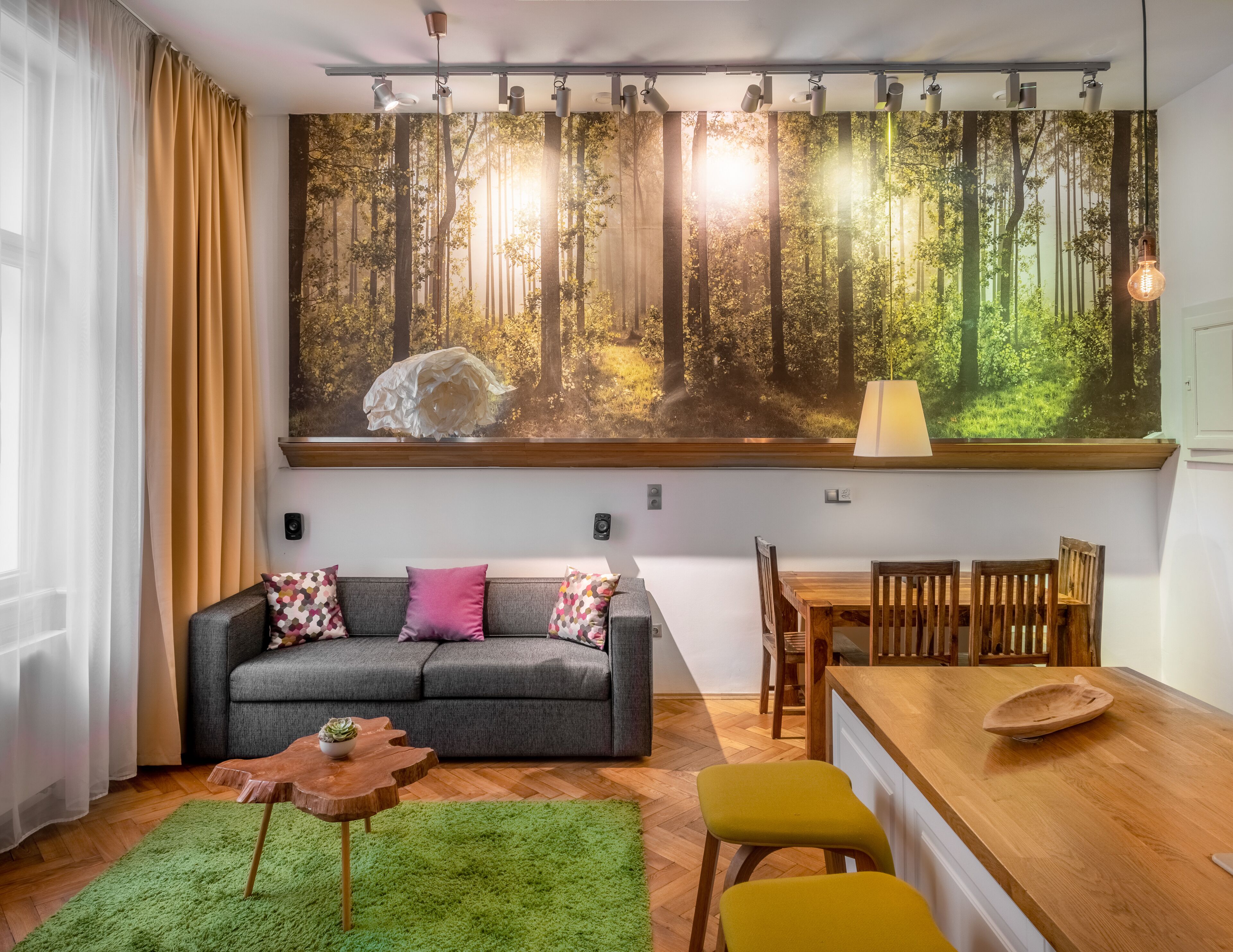 A living room with a grey sofa, colorful cushions, a wooden coffee table, a dining table with wooden chairs, and a large forest mural on the wall.