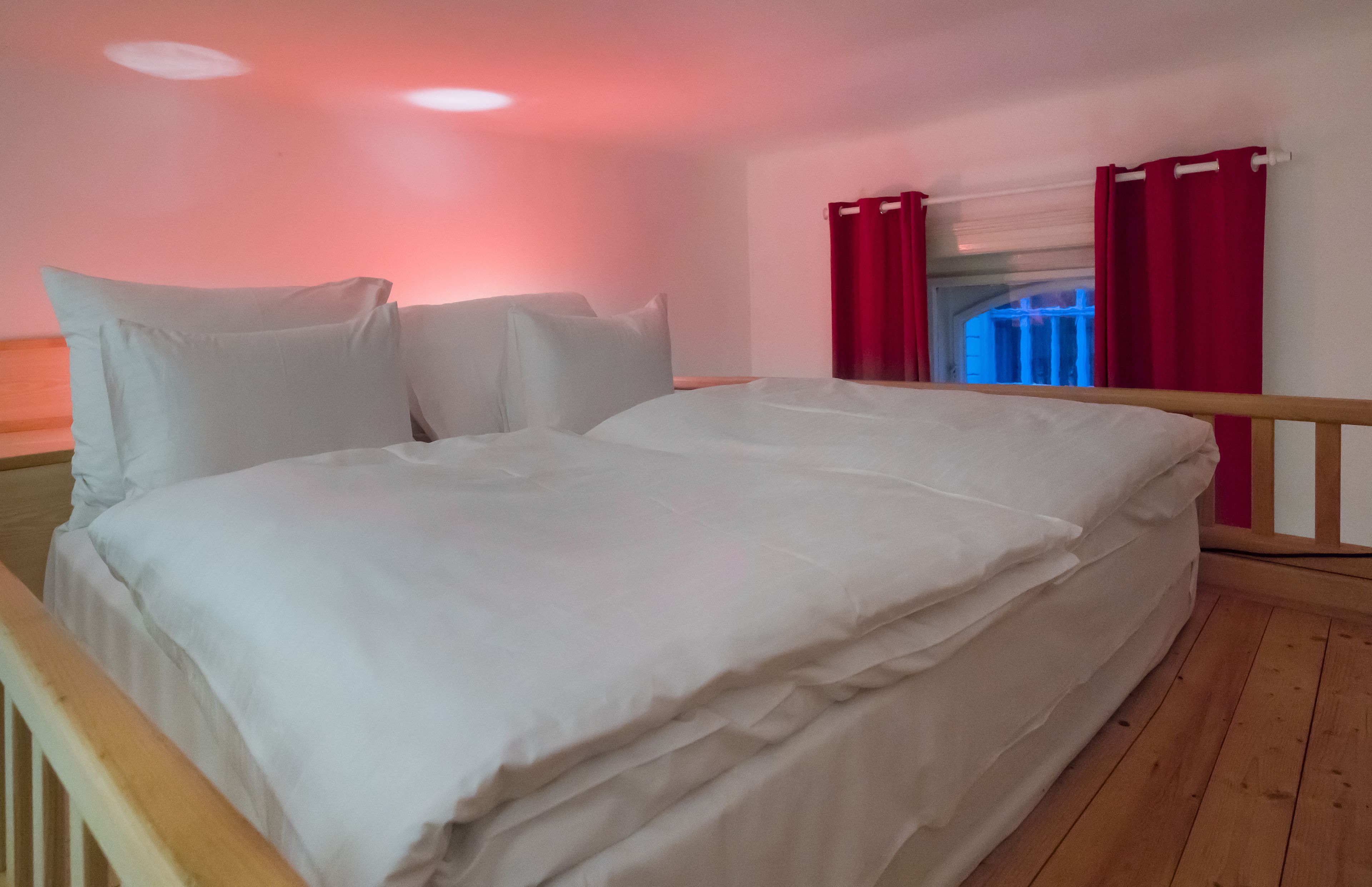 Un lit bien fait, avec du linge de lit blanc, dans une pièce au plancher en bois et aux rideaux rouges.