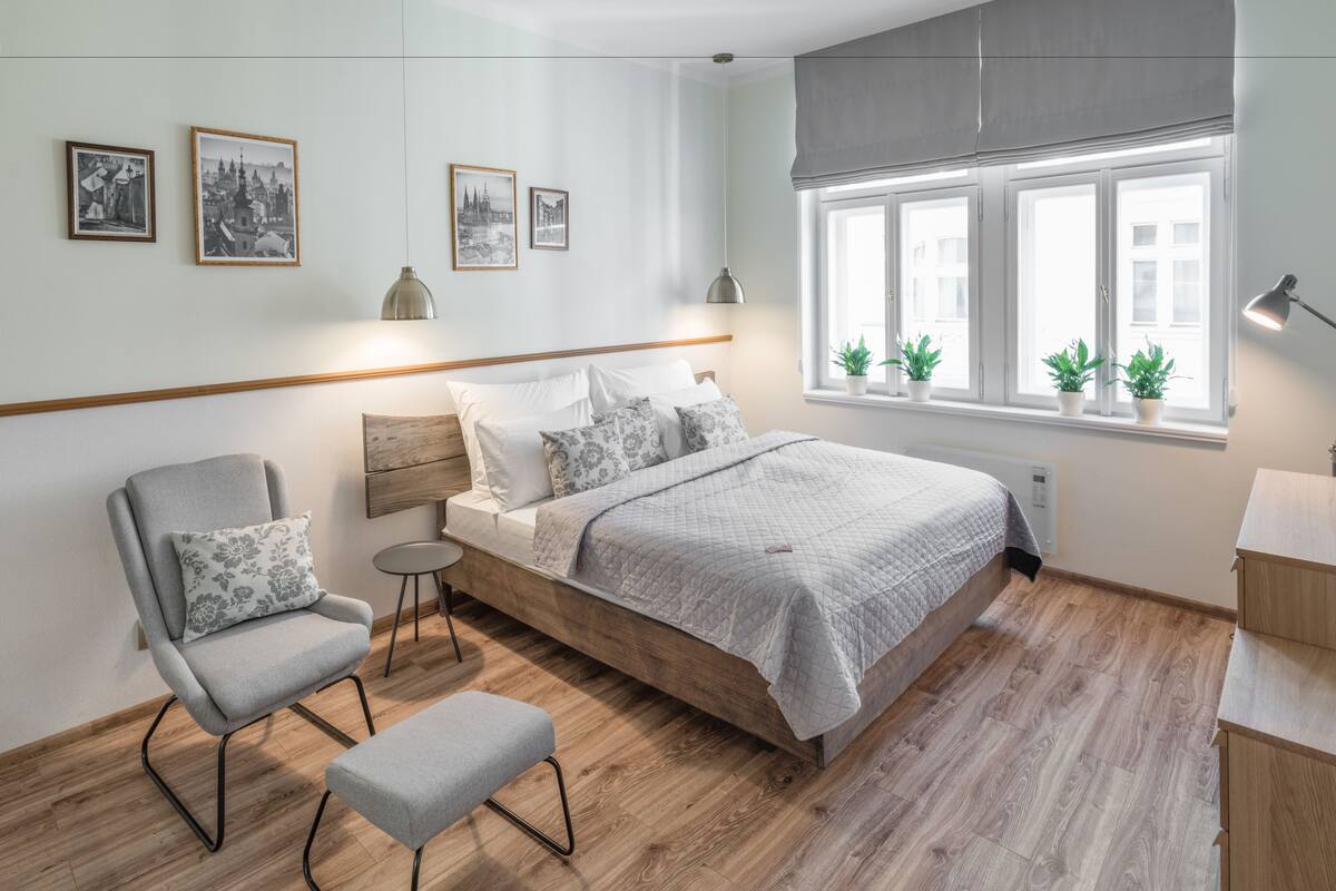 A modern bedroom with a wooden bed, a grey armchair, a small round table, and a bench. There are three framed pictures on the wall, a window with plants, and a bedside lamp.