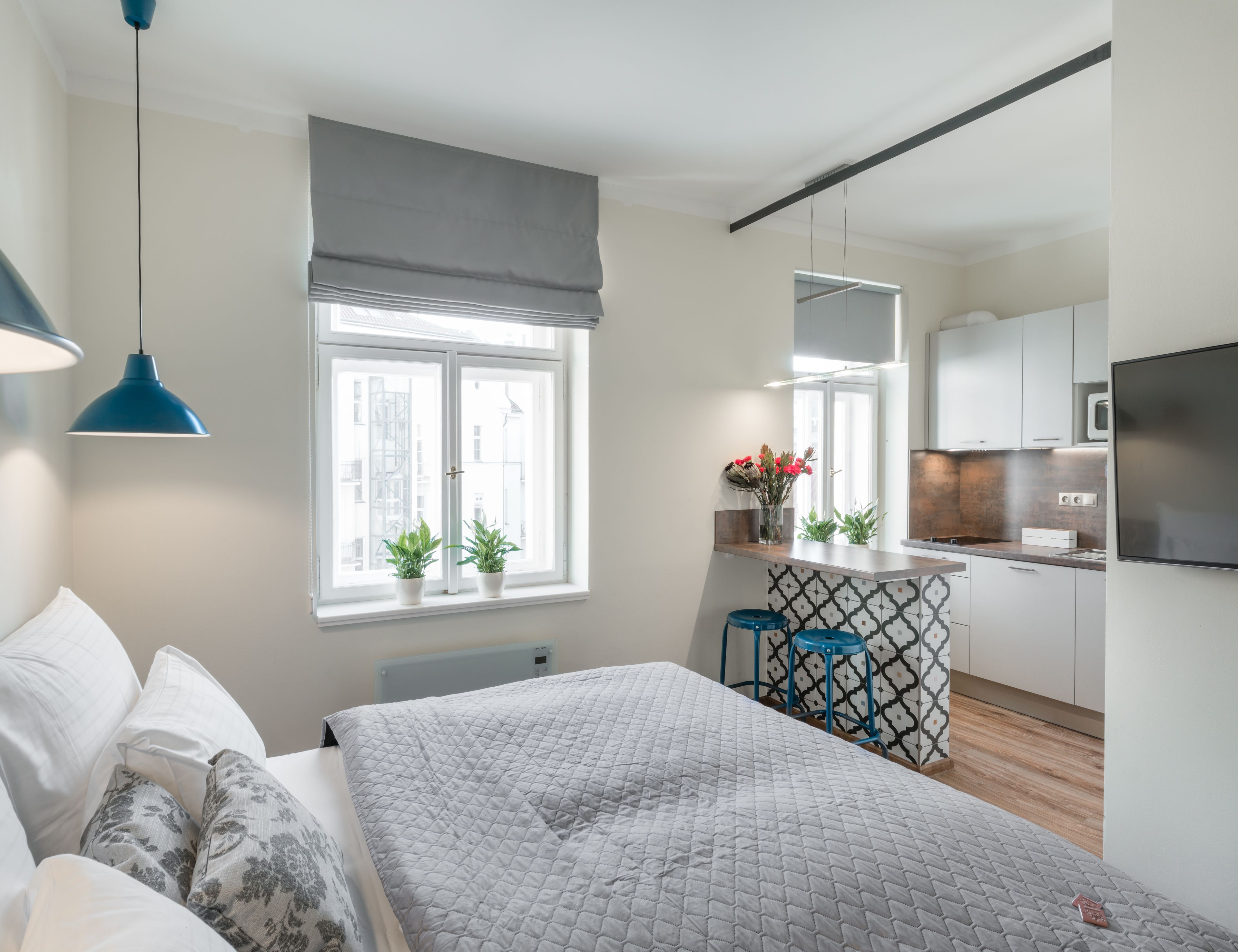A modern bedroom with a large bed, a bedside table, a window with a view of buildings, a kitchen area with a microwave, and a blue pendant light.