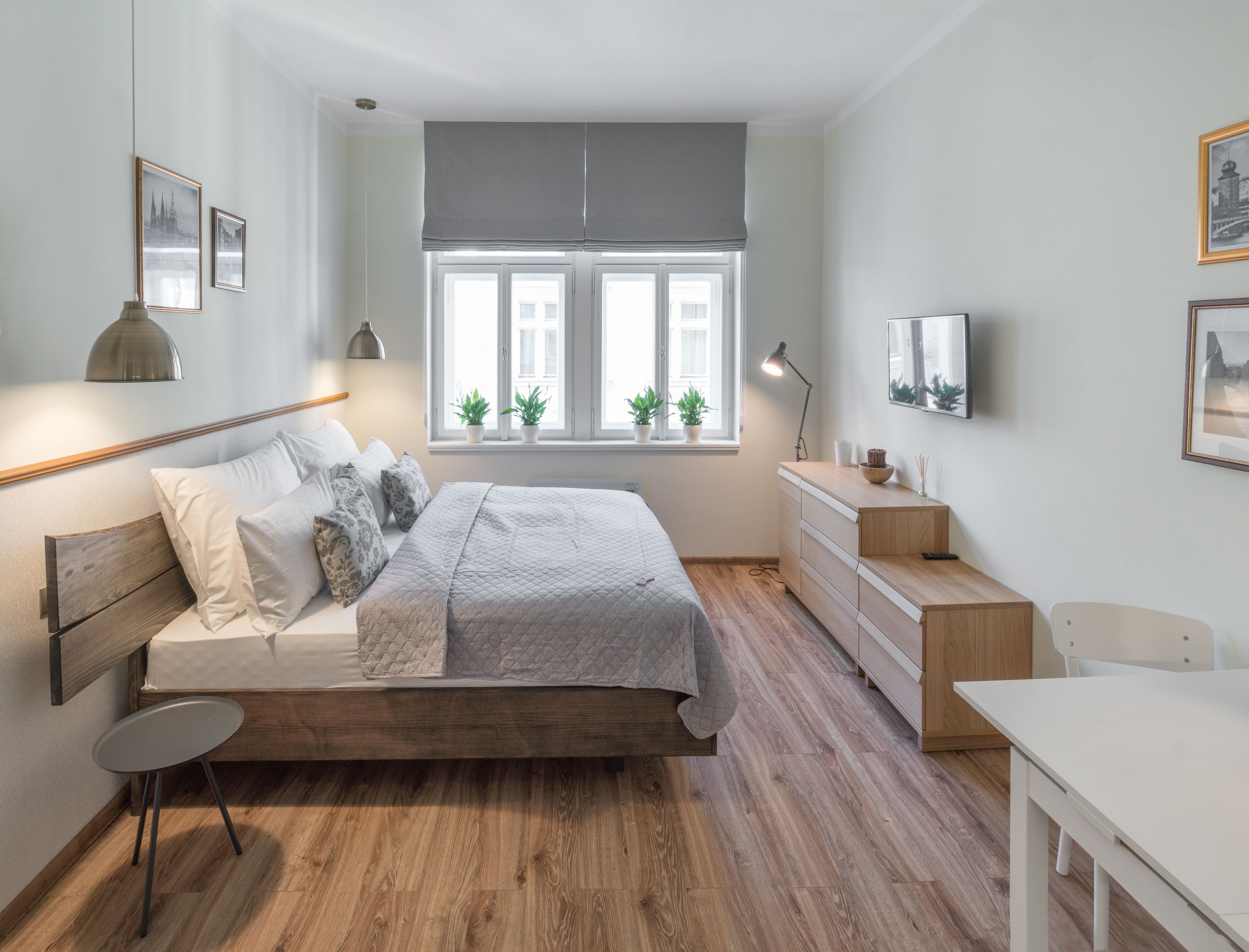 A modern bedroom with a wooden bed, bedside table, and a desk. There are two windows with plants on the windowsill, and a ceiling fan above.