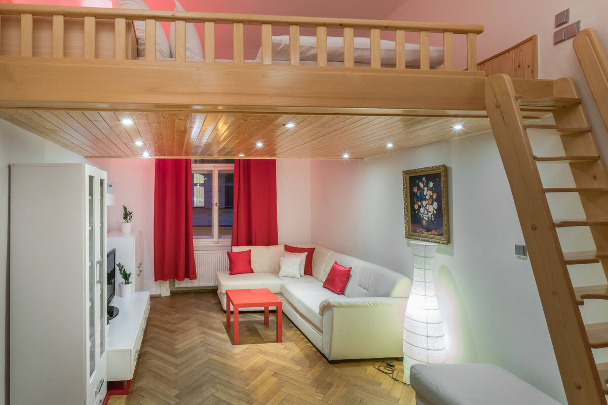 A loft-style room with a wooden ceiling, white sofa, red cushions, a small table, and a ladder.