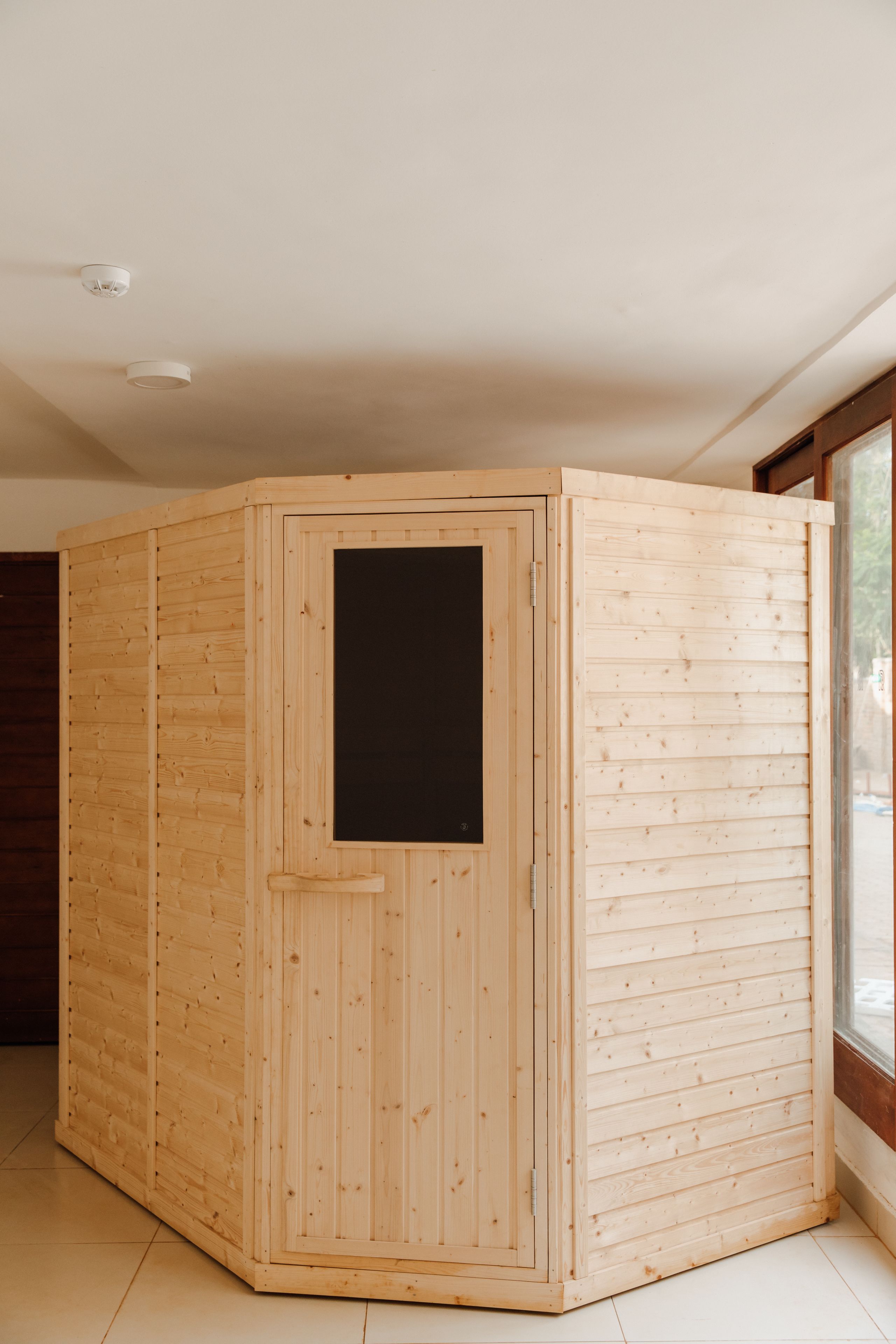 A wooden sauna with a sliding door, located in a room with a large window.