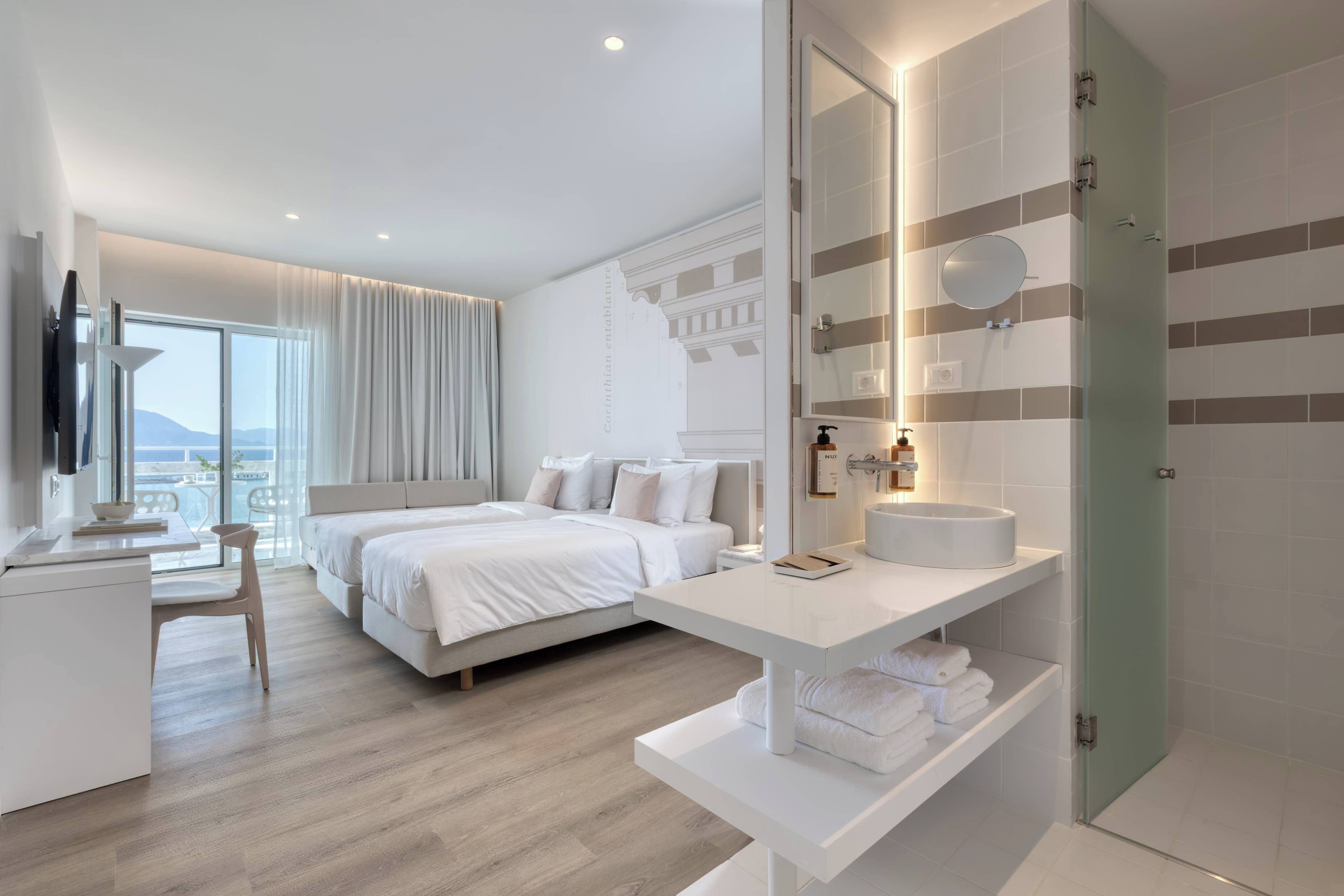 A modern hotel room with a large bed, a desk, a bathroom with a mirror, and a view of the sea.