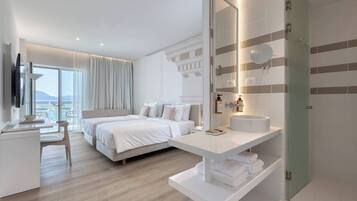 A modern hotel room with a large bed, a desk, a bathroom with a mirror, and a view of the sea.