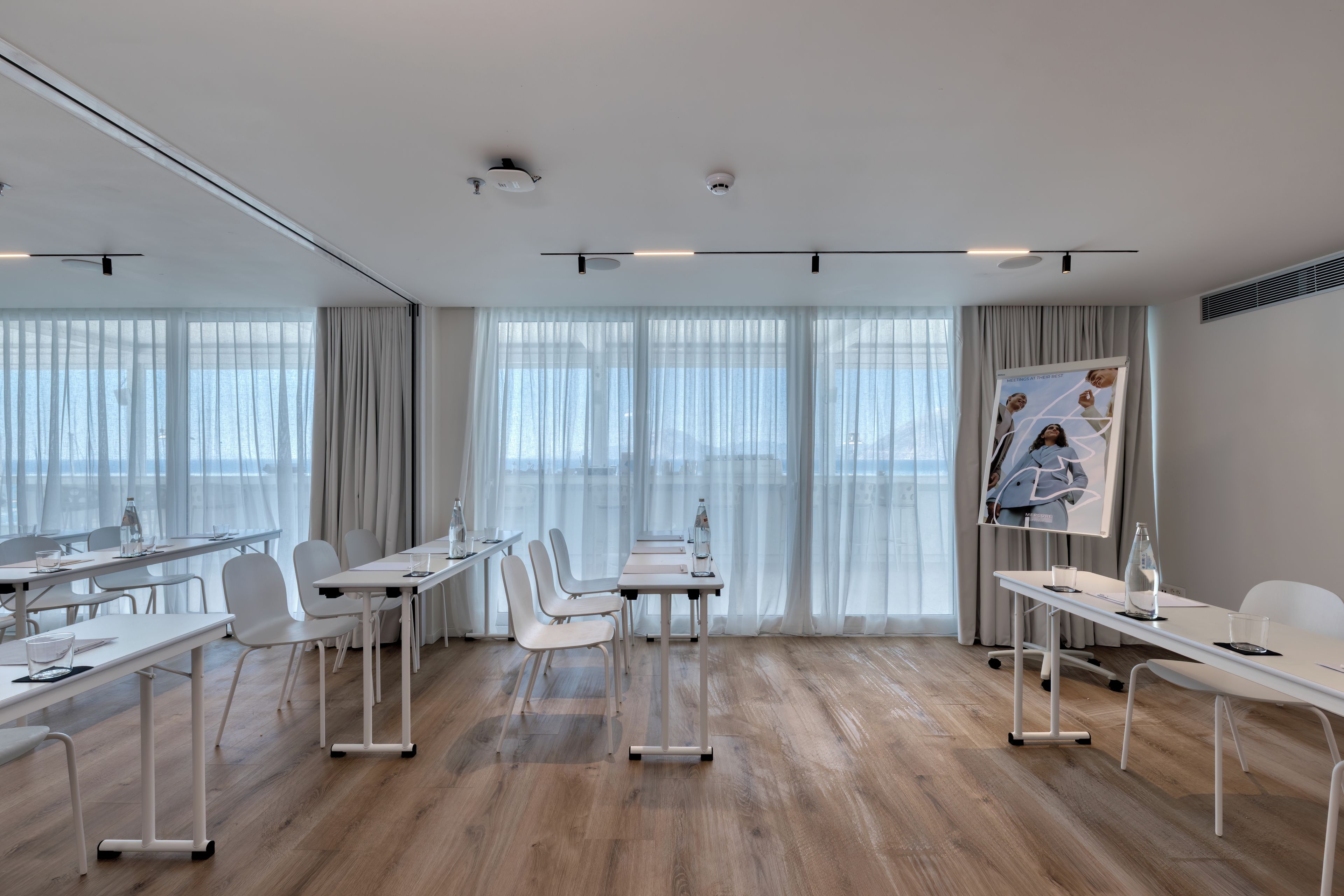 A modern conference room with white furniture, large windows with sheer curtains, and a poster on the wall.