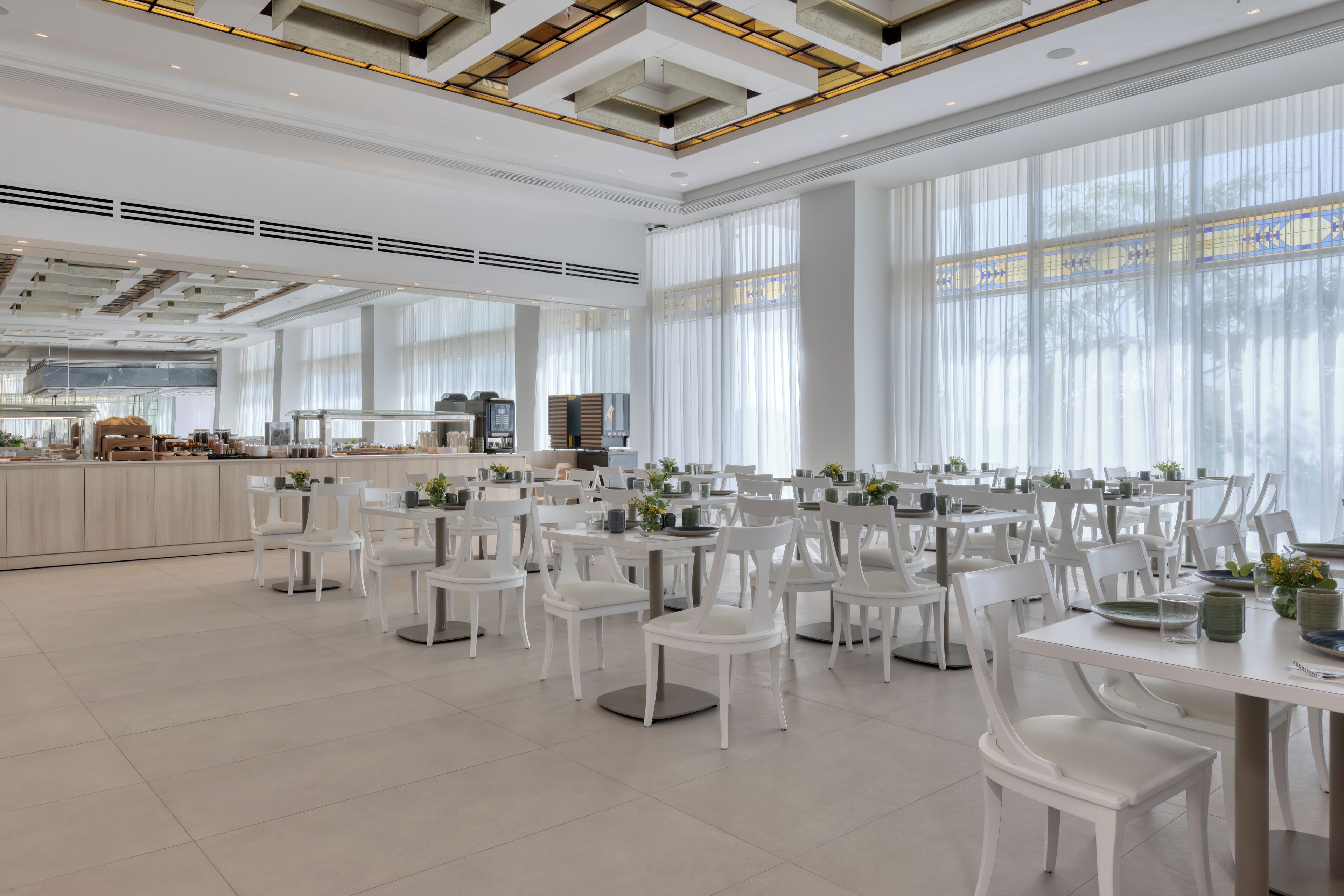 A spacious dining area with white chairs and tables, large windows, and a modern ceiling design.