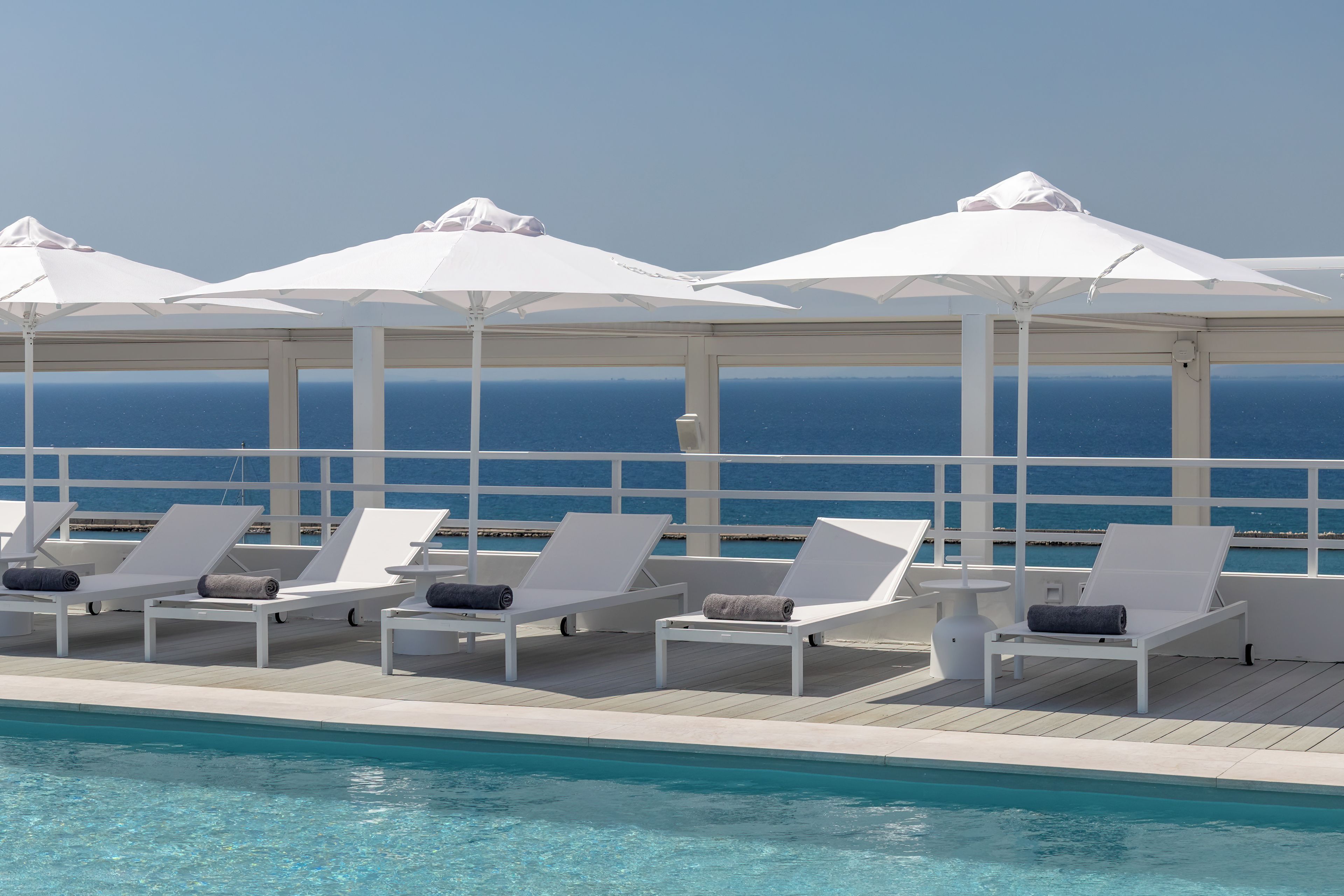 Zona de piscina con sillas reclinables blancas y sombrillas, con vista al mar azul cristalino.