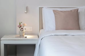 A neatly made bed with white bedding and a single beige pillow, a bedside table with a vase of flowers, and a wall-mounted light. - Mercure Patras Astir (Patras)
