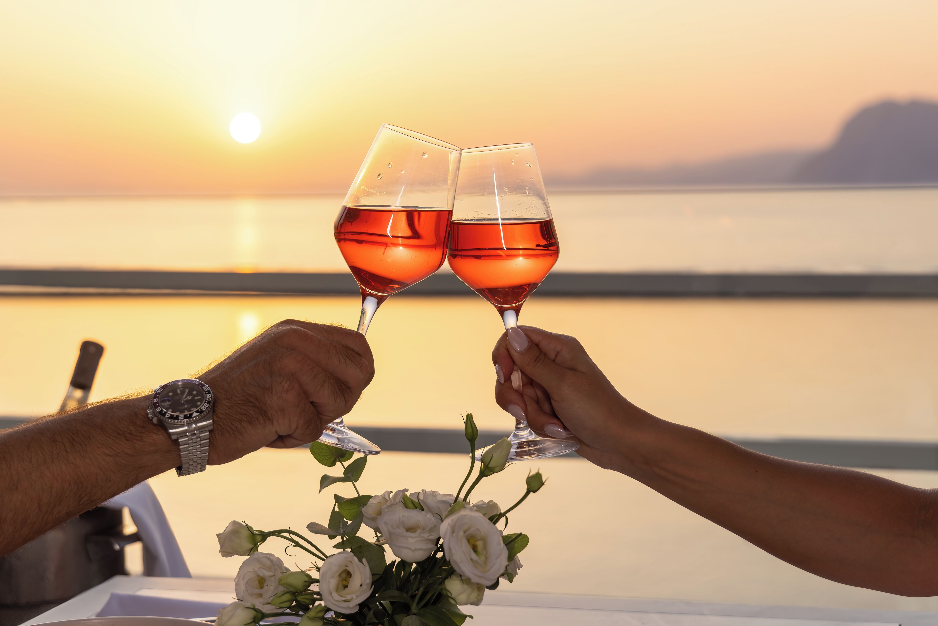 Two people toasting with wine glasses, a bouquet of white flowers, and a sunset view.