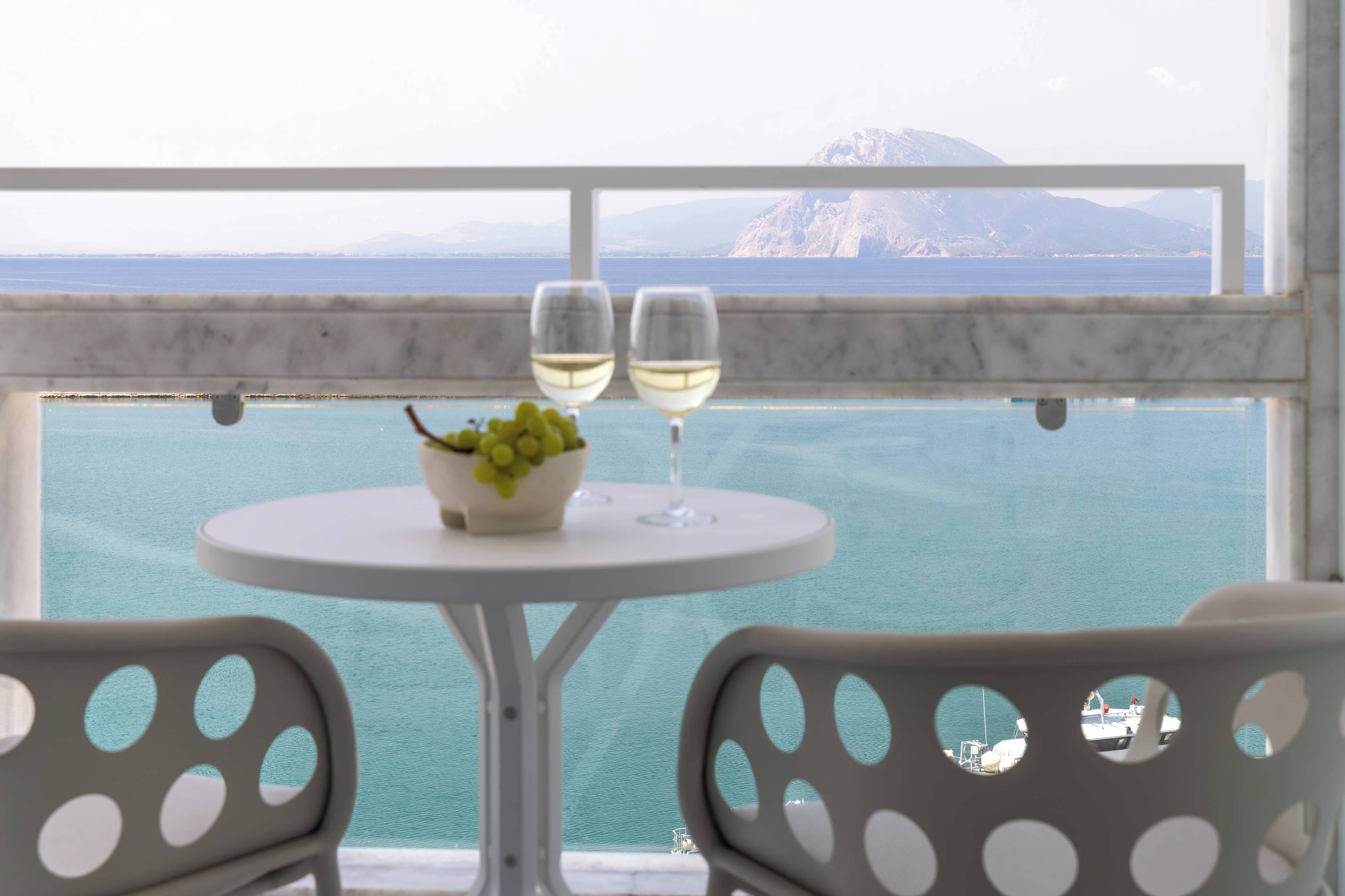 Uma mesa com uma tigela de uvas e dois copos de vinho branco, com vista para a água e uma montanha.