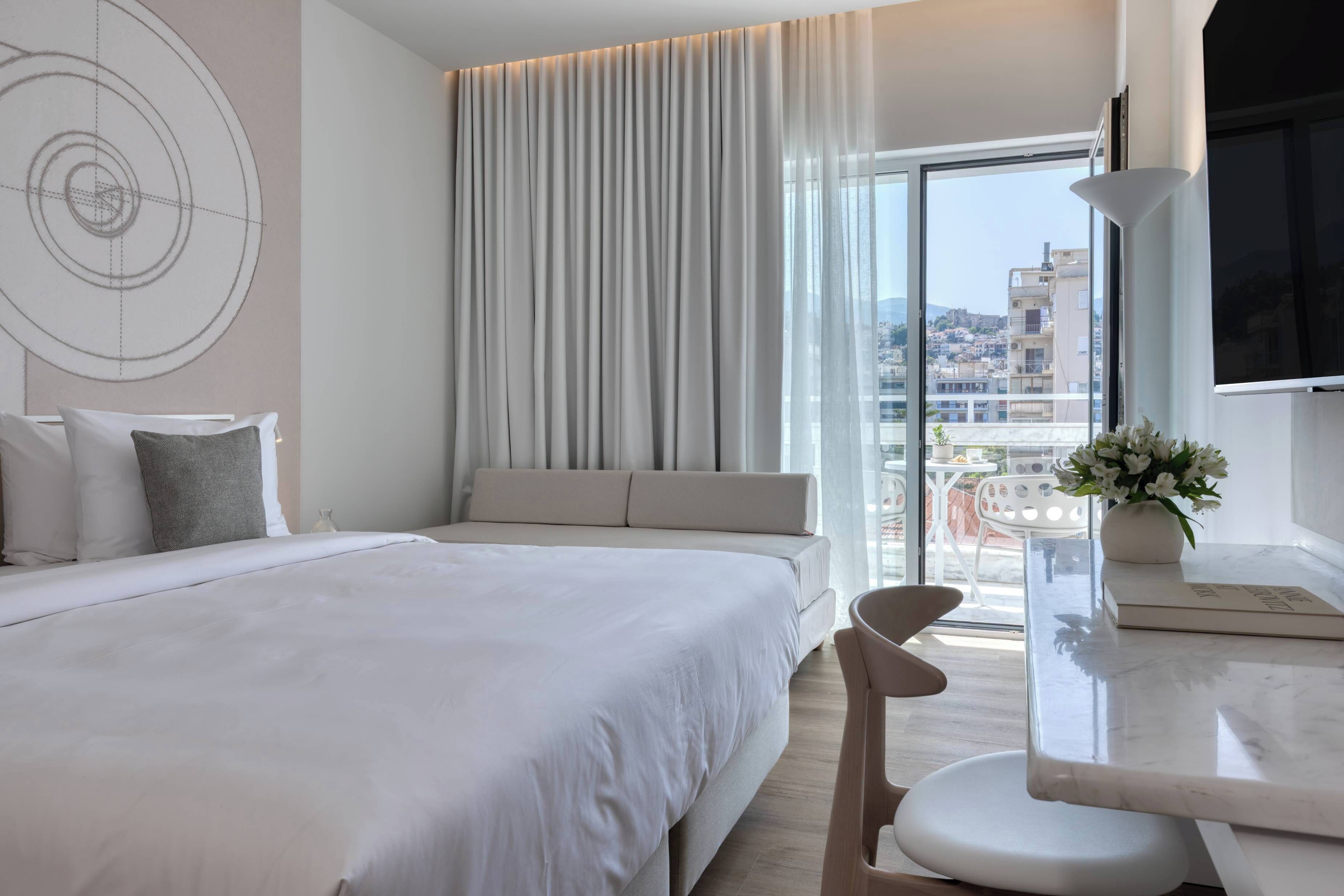 Ein Hotelzimmer mit Bett, einer Couch, einem Fernseher, einem Tisch mit Blumenvase und Blick auf die Stadt.