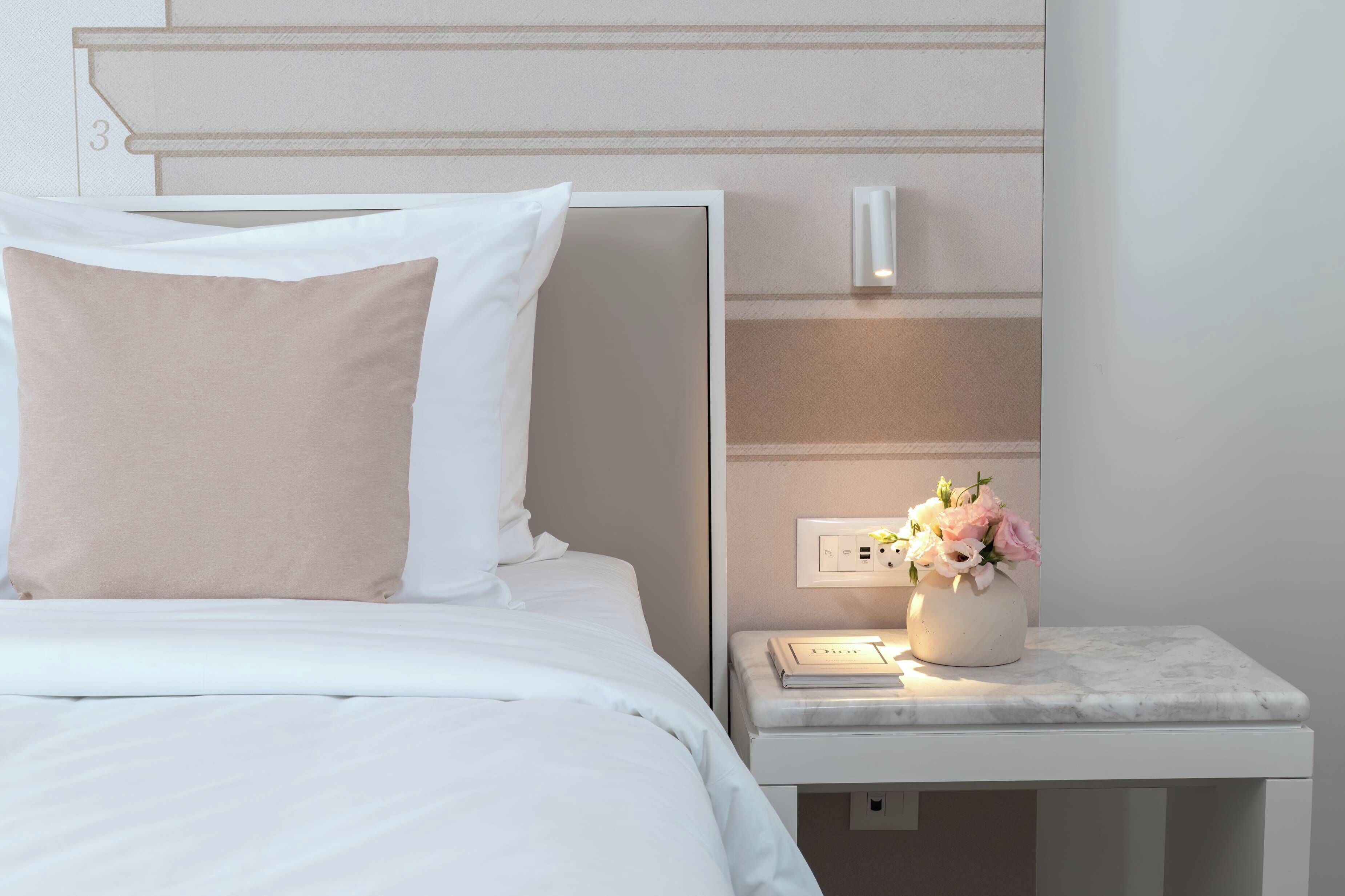 A neatly made bed with white bedding and a beige pillow, a bedside table with a vase of flowers, and a wall-mounted light fixture.