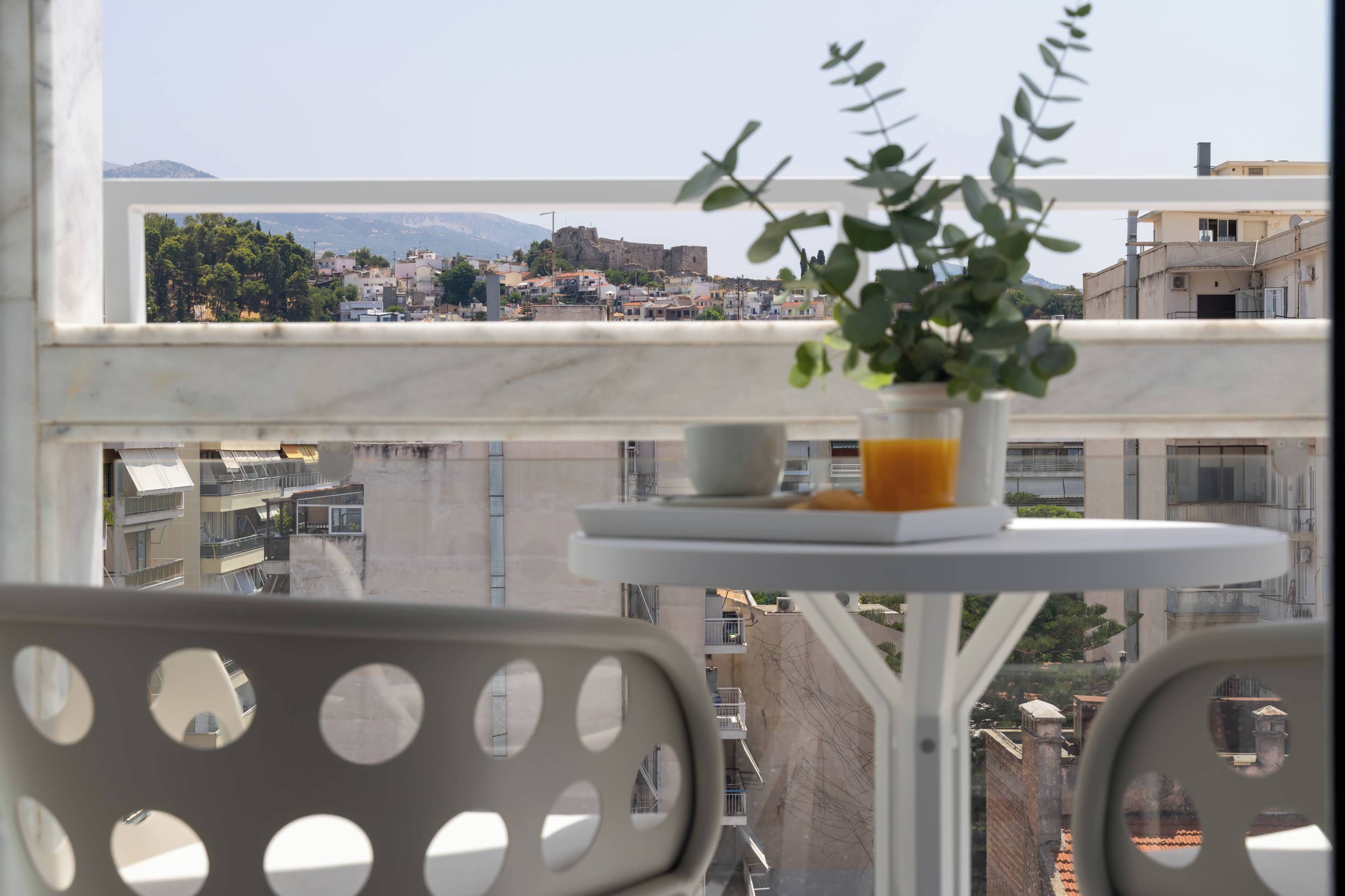 Un balcón con planta en maceta, una mesa y vistas de un paisaje urbano con edificios y una montaña.
