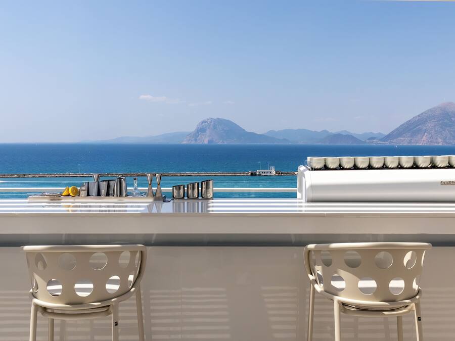 A view from a balcony with two chairs, a table, and a bar area overlooking the sea and distant mountains.