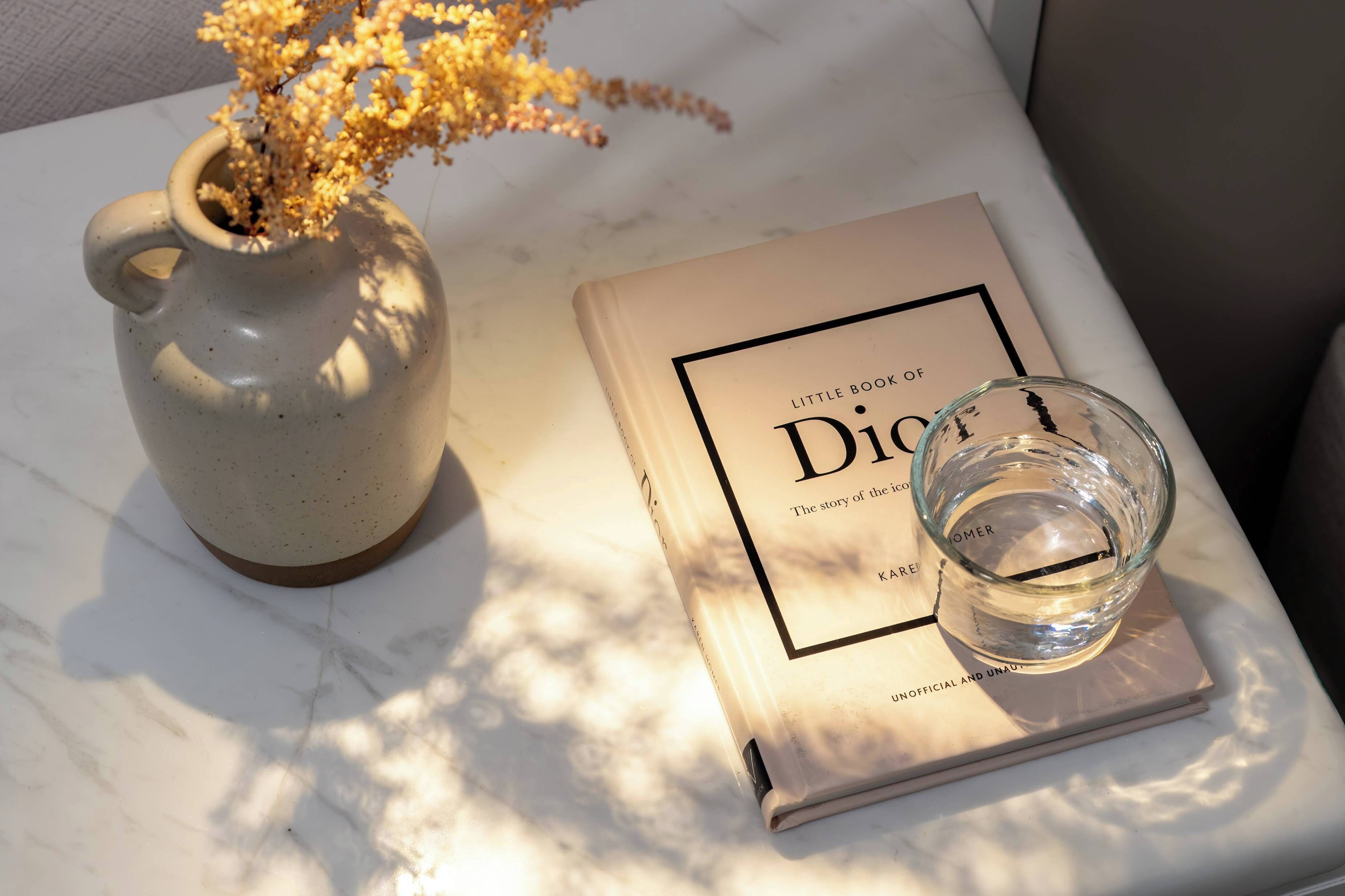 A vase with dried flowers, a book titled 'Little Book of Dior,' and a clear glass on a marble surface.