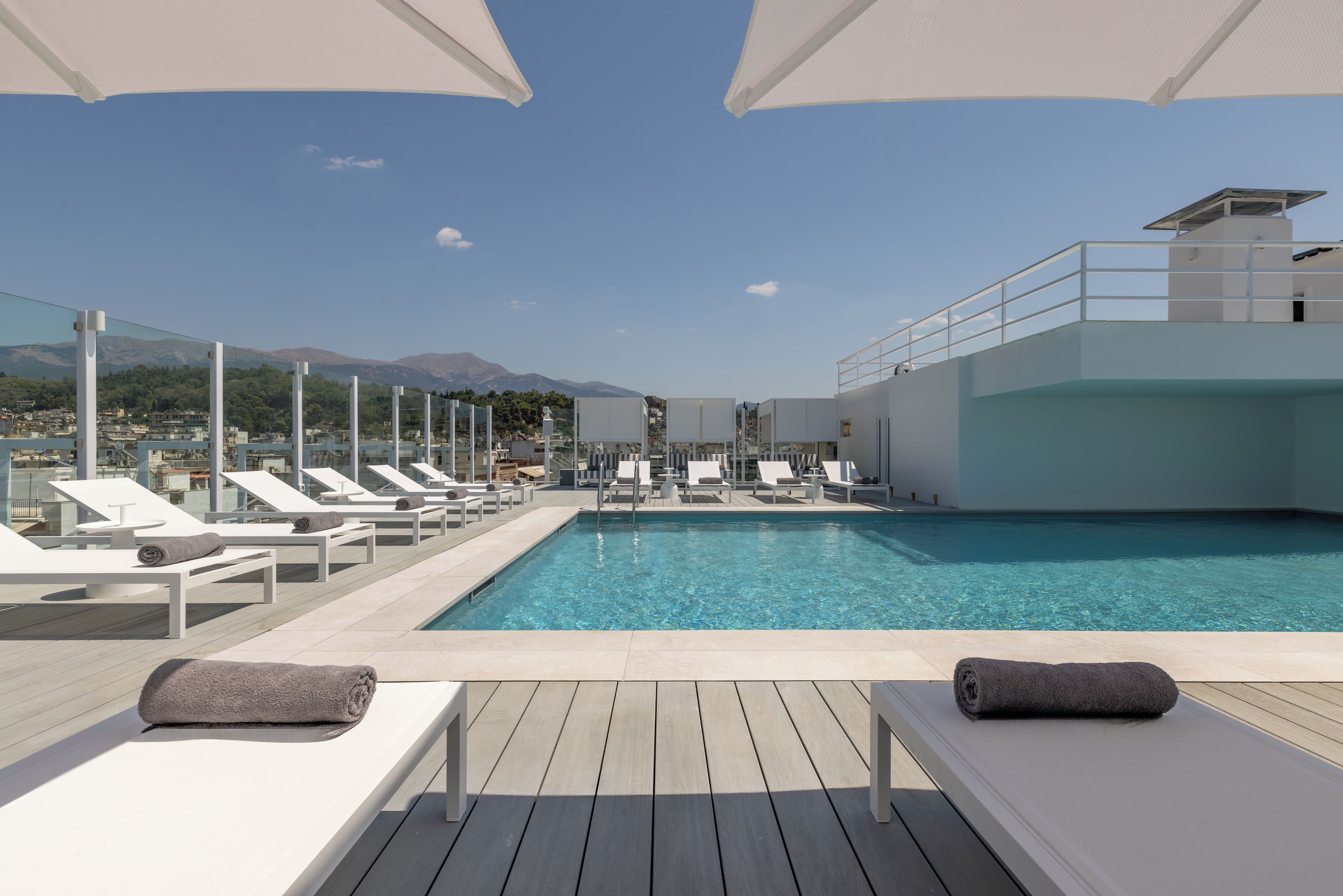 A rooftop pool with lounge chairs and umbrellas, overlooking a mountainous landscape.