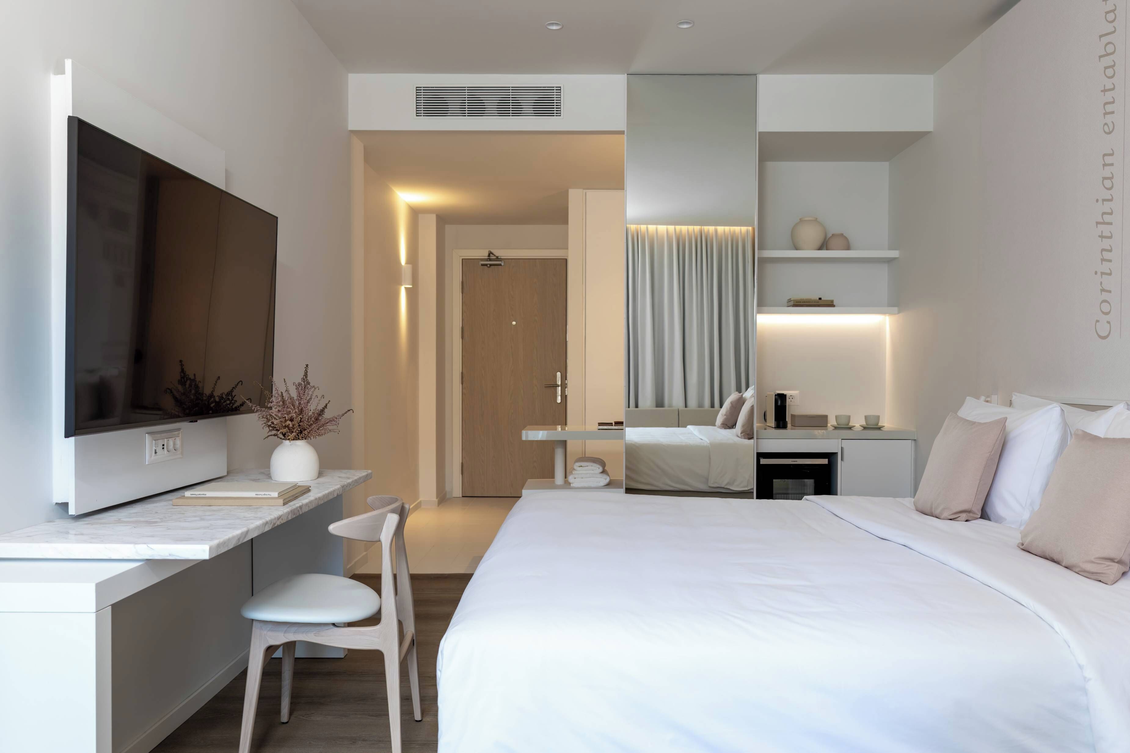 A modern hotel room with a large bed, a desk with a flat-screen TV, a seating area with a chair, a vanity with a mirror, and a kitchenette.