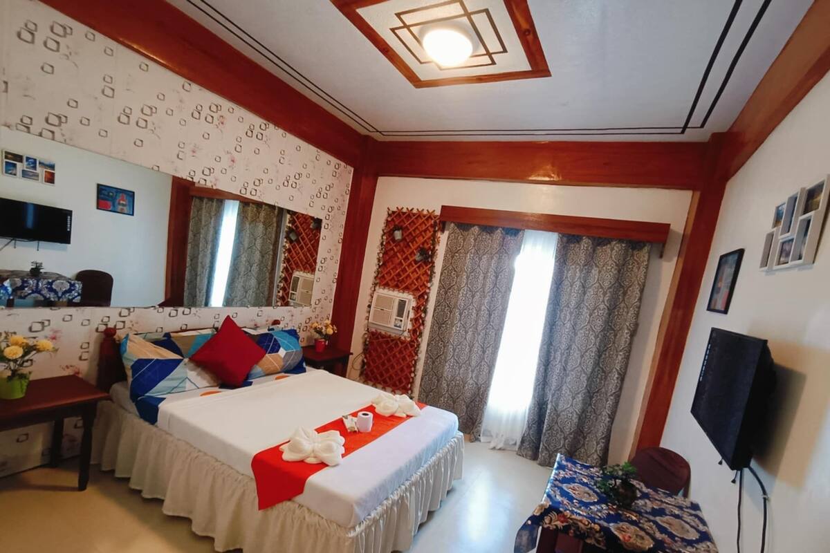 A bedroom with a bed, a TV, a desk, and a window with curtains.