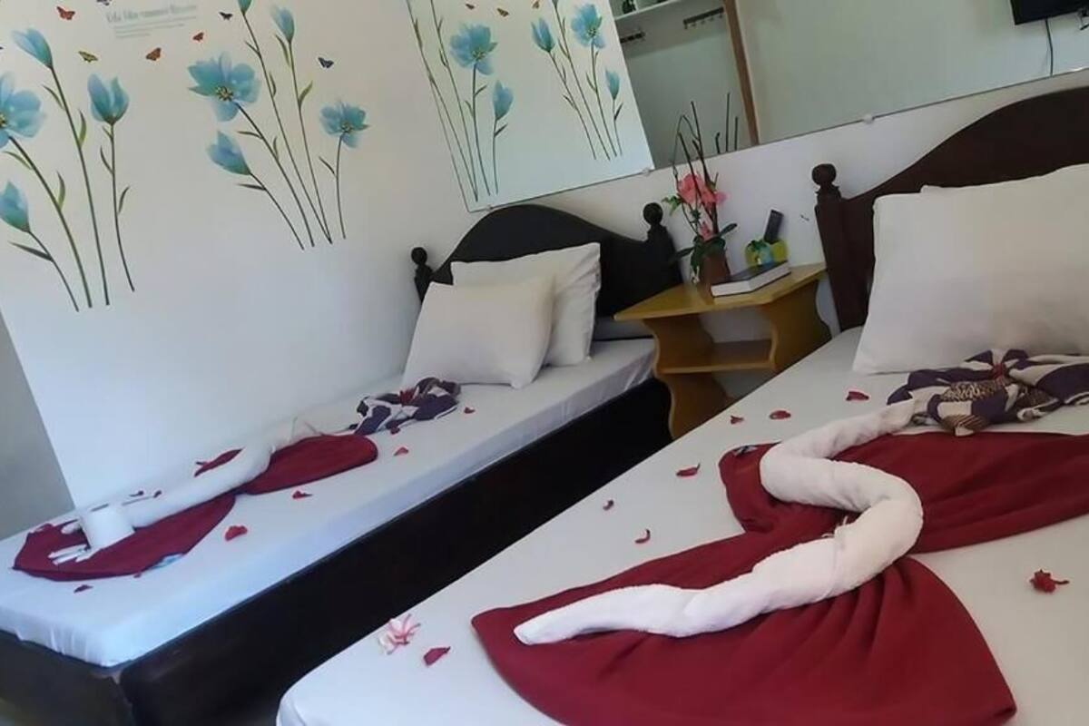 A hotel room with two beds, each with a white pillow and a red blanket. There is a wooden nightstand with a lamp and a vase of flowers. The room has a large mirror on the wall and a floral wall decoration.