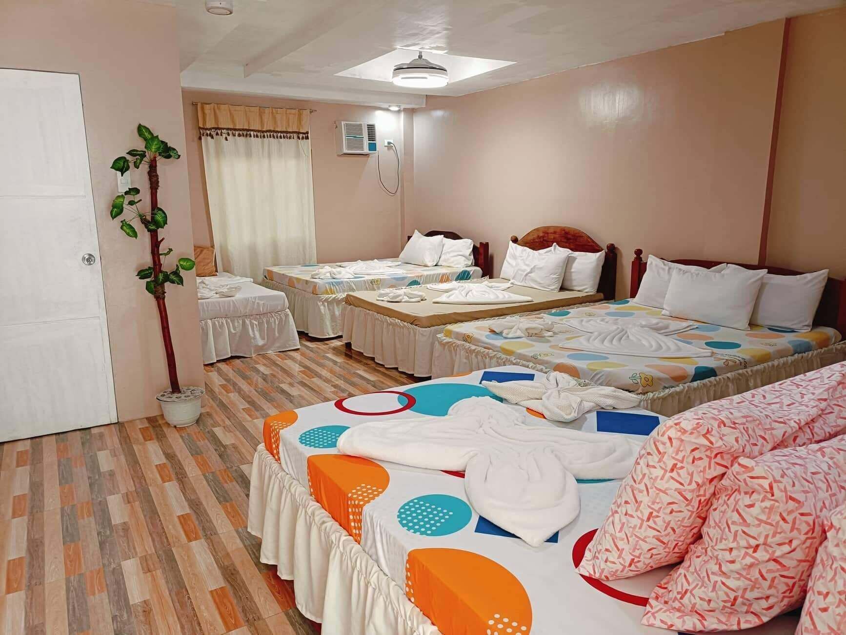 Habitación de hotel con dos camas individuales, una cama doble, una planta en maceta y un ventilador de techo.
