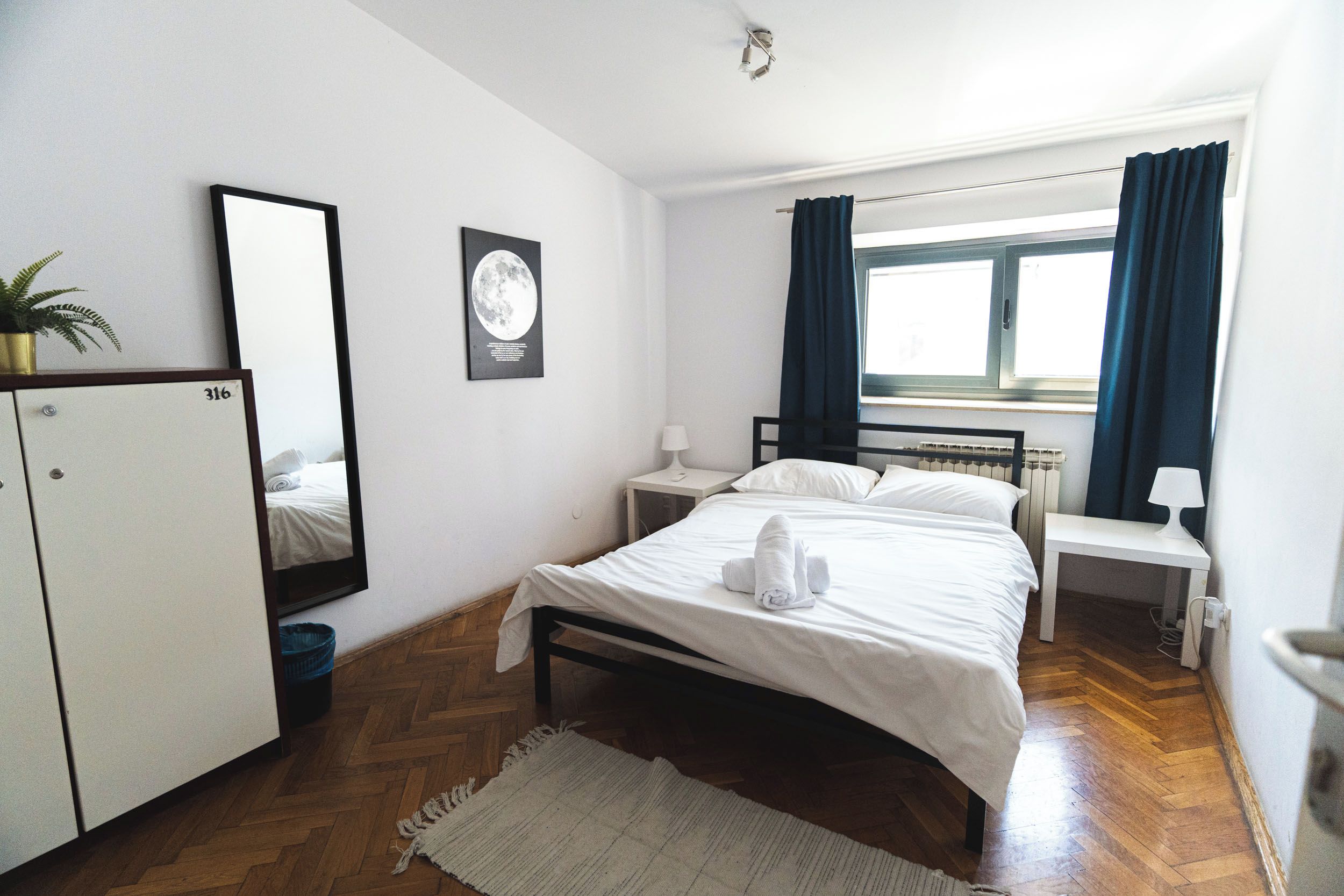 A hotel room with a bed, a nightstand, a desk, a wardrobe, a window with curtains, and a wall-mounted mirror.