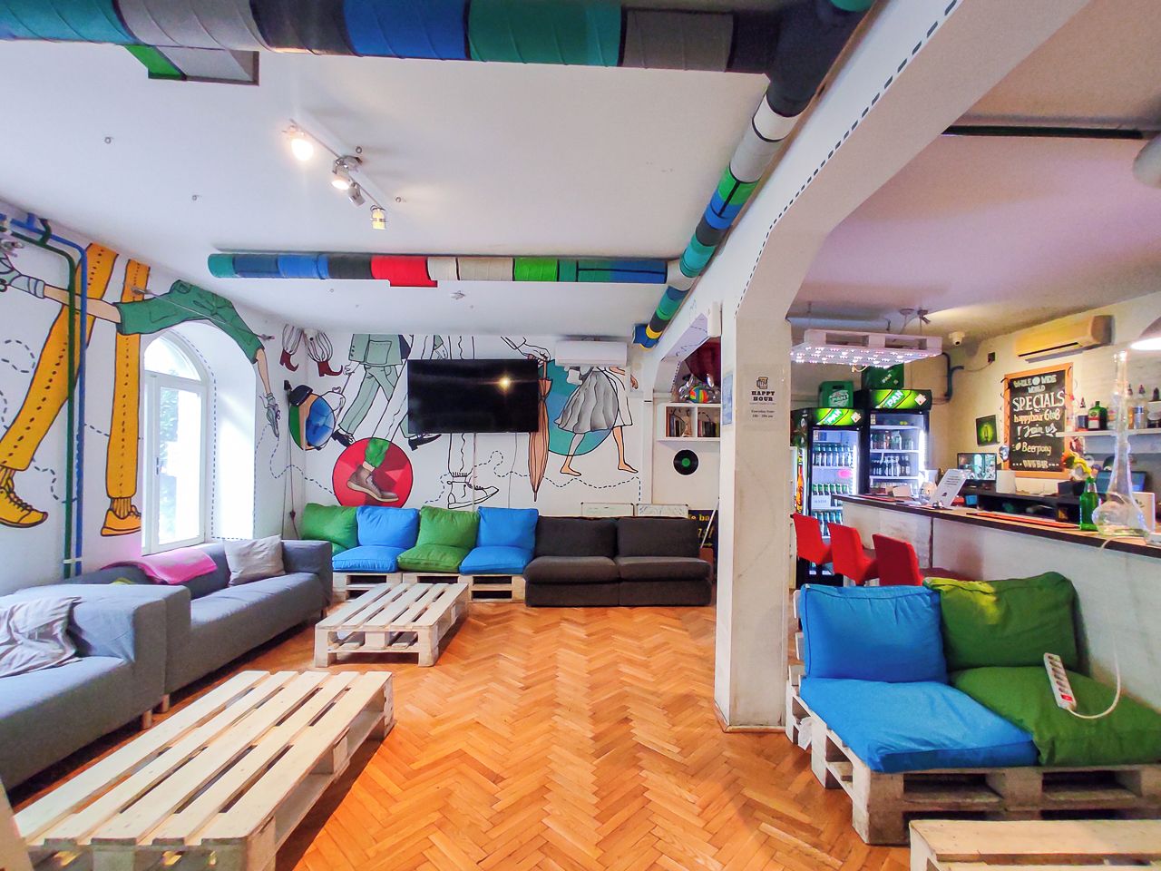 A modern living room with a herringbone floor, a flat-screen TV mounted on the wall, and a bar area with a neon sign.