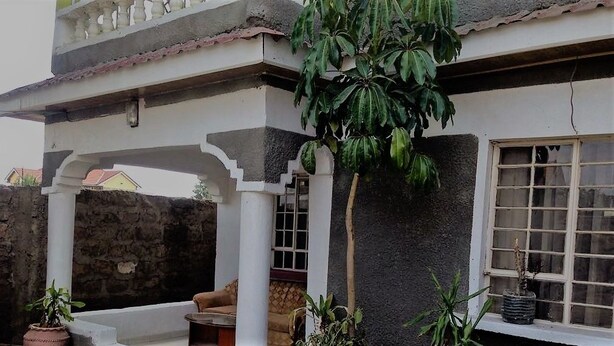 A covered outdoor area with a stone wall, a white column, and a window with a plant on the sill. - Nairobi Transit Lounge (Syokimau)