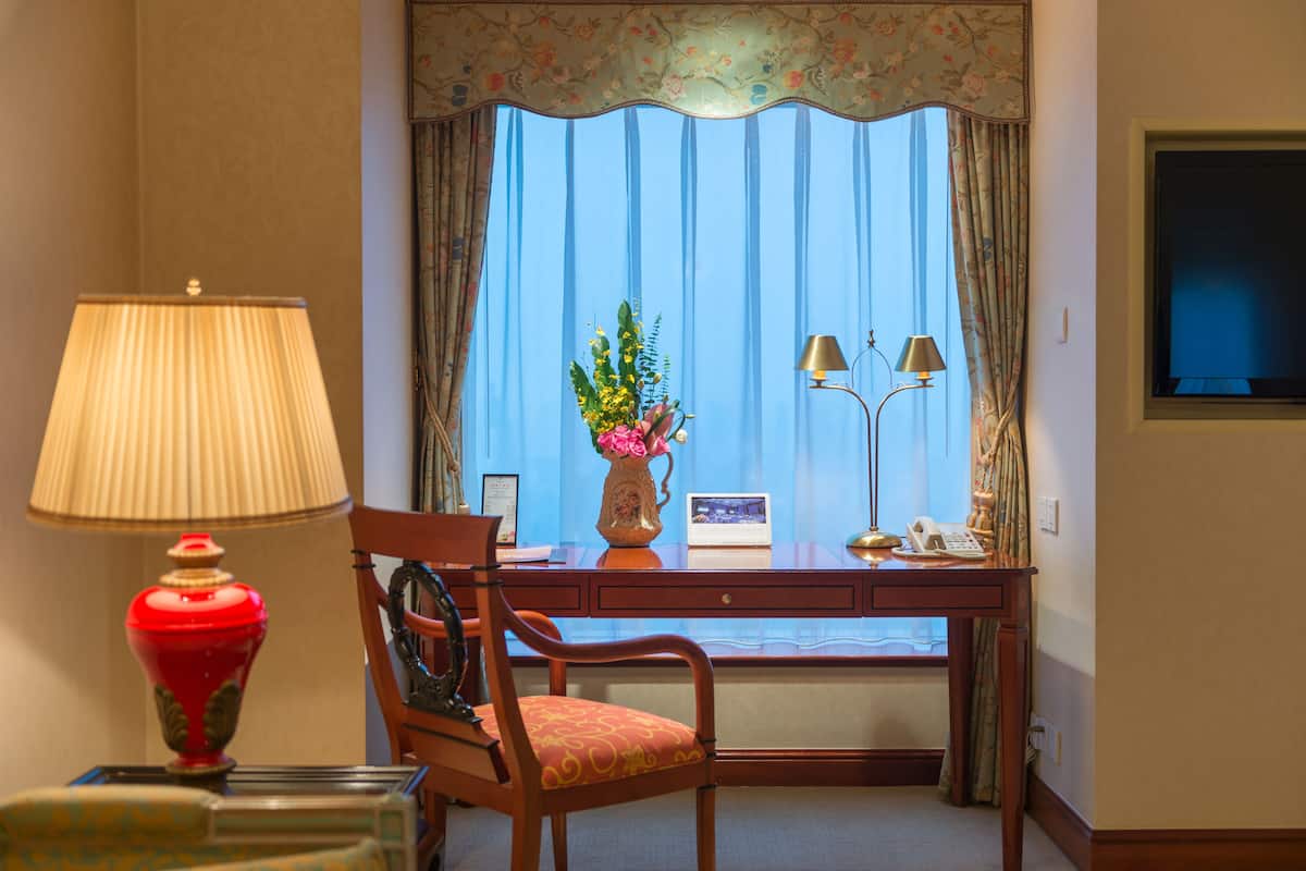 Habitación de hotel con escritorio, silla, lámpara y ventana con cortinas.