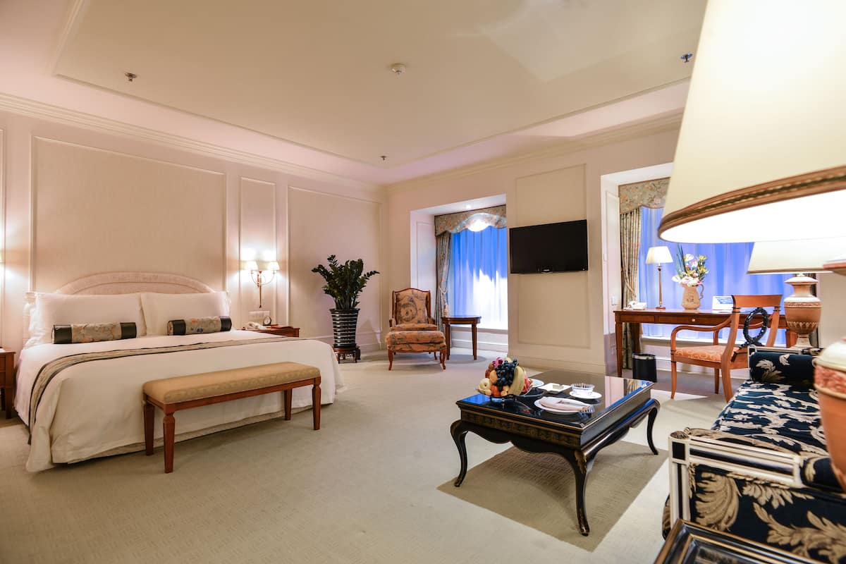 Una habitación de hotel con una cama grande, un área de descanso con sofá y sillones, una mesa de comedor y un televisor.
