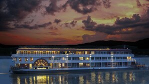 Un gran barco de vapor fluvial con varias cubiertas y una imponente casa de máquinas, atracado al atardecer con reflejos en el agua.