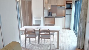 A modern living room with a sofa, a dining table with chairs, a kitchen area with cabinets and appliances, and a large window with curtains. - Terres de France - Domaine du Moulin Neuf (Malansac)