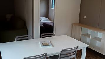 Habitación compacta con una mesa blanca y dos sillas, una cama junto a una ventana y un mueble empotrado para almacenaje.