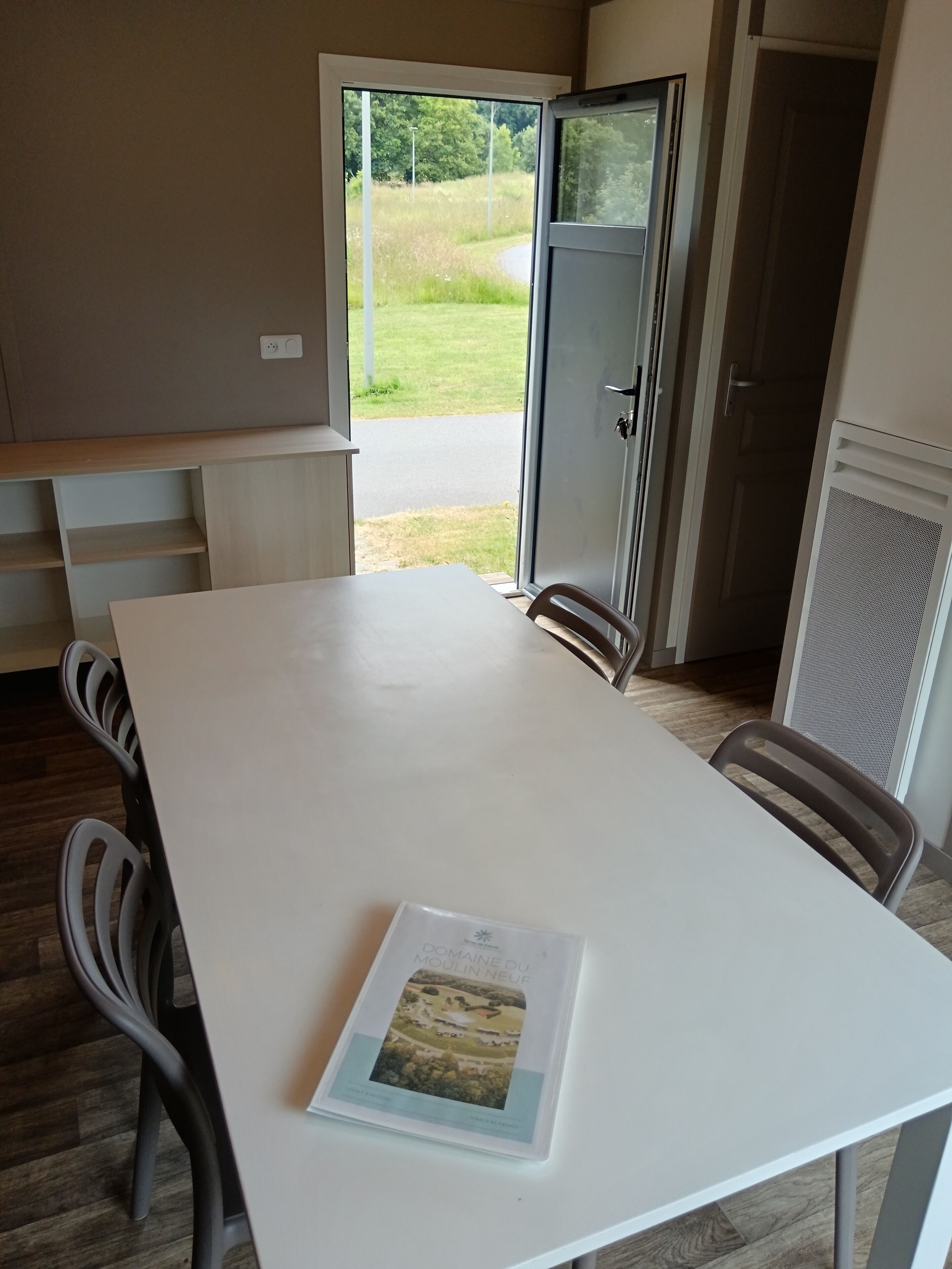 Un espace repas moderne avec une table blanche, deux chaises et une vue sur un paysage verdoyant, visible à travers une porte ouverte.