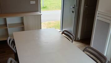 Un espace repas moderne avec une table blanche, deux chaises et une vue sur un paysage verdoyant, visible à travers une porte ouverte.