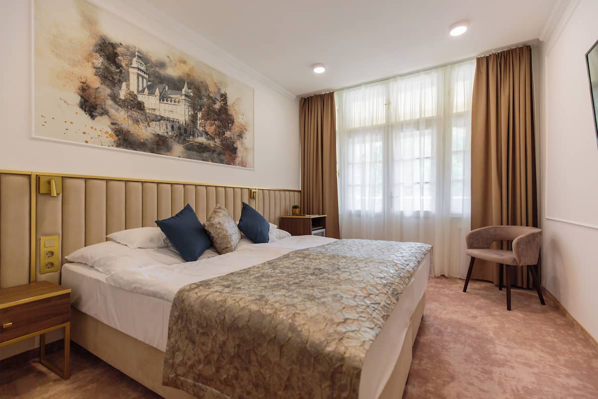 A hotel room with a large bed, a nightstand, a chair, and a painting on the wall.