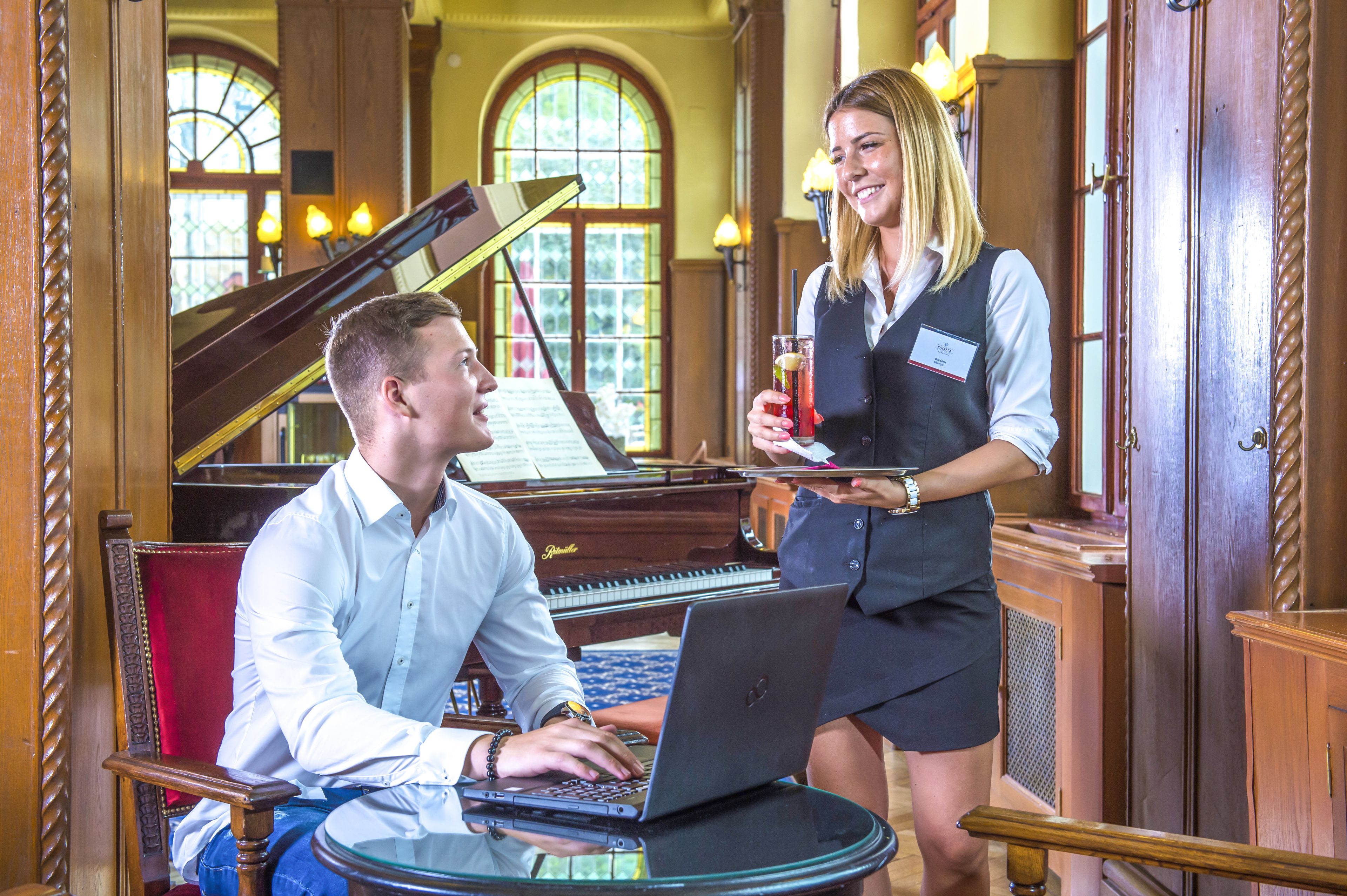 A man seated at a table with a laptop, a grand piano in the background, and a woman standing beside him holding a drink.
