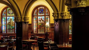 A dining area with high arched windows and stained glass, wooden tables and chairs, and a chandelier. - Hotel Palota Lillafüred (Miskolc)