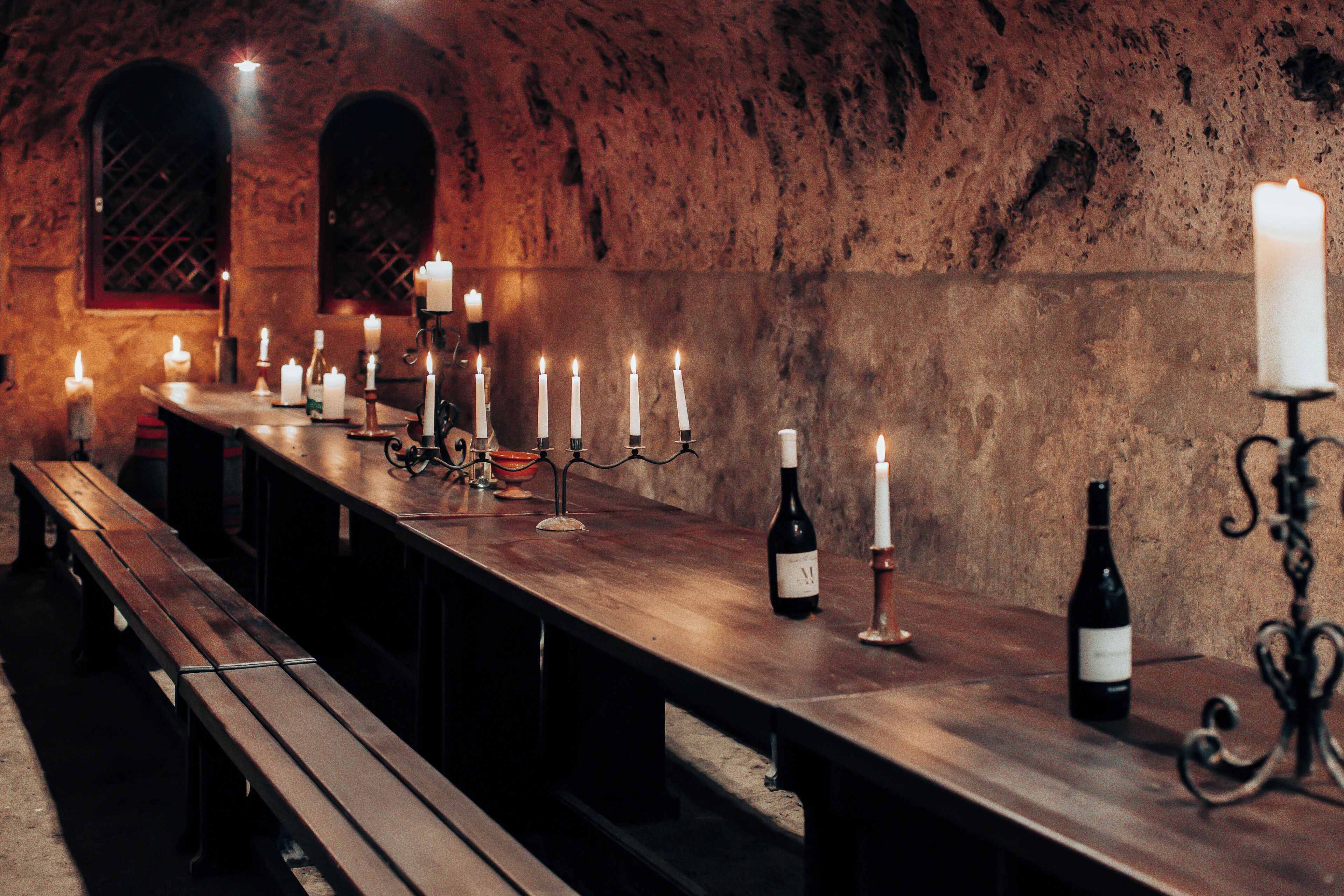 A long wooden table with candles and wine bottles, set against a stone wall with arched windows.
