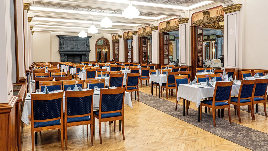 A large dining hall with rows of tables and chairs, a fireplace, and ornate wall decorations.